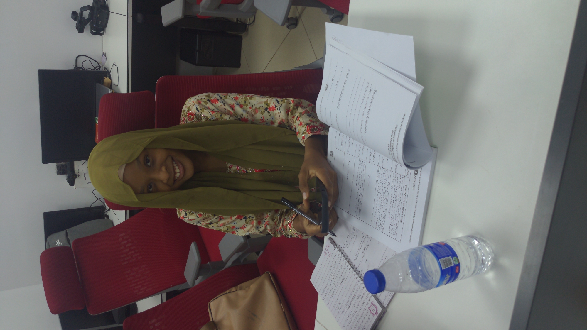 This media file captures a smiling young woman seated at a desk in what appears to be a modern office or study environment, likely in Abuja, Nigeria. The image is rotated 90 degrees clockwise.
The woman, likely in her late teens or early twenties, is wearing a muted olive-green head covering (khimar or hijab) and a light-colored, long-sleeved floral top. She looks directly at the camera with a broad, friendly smile.
She appears to be engaged in an activity related to work or study. Her right hand holds a black pen, poised over an open notebook or document on the white desk, while her left hand holds a black smartphone or similar device. A stack of papers with printed text is visible on the desk, along with a clear plastic water bottle with a blue cap and blue label, marked "SA" and "AQUA".
The desk area is equipped with a computer monitor, a video camera or webcam on a tripod, and various cables. Two red office chairs are visible, one occupied by the woman and another partially visible behind her. A brown bag or purse rests on the floor next to her chair. Underneath a monitor, a grey laptop bag with the partial text "gle" (likely "Google") is visible.
Visible text on the documents includes phrases such as "Summary of weekly mot", "New knowledge", "Student", "ing the Gap Between Theory & Practice Creativity Innovation & Productivity", "4 ACTIVITIES", "TIME", "DESCRIPTION OF WORK DONE", and days of the week like "WEDNESDAY", "SATURDAY", and partially "MONDAY". There are also handwritten notes on the pages. The woman's expression conveys happiness and engagement.