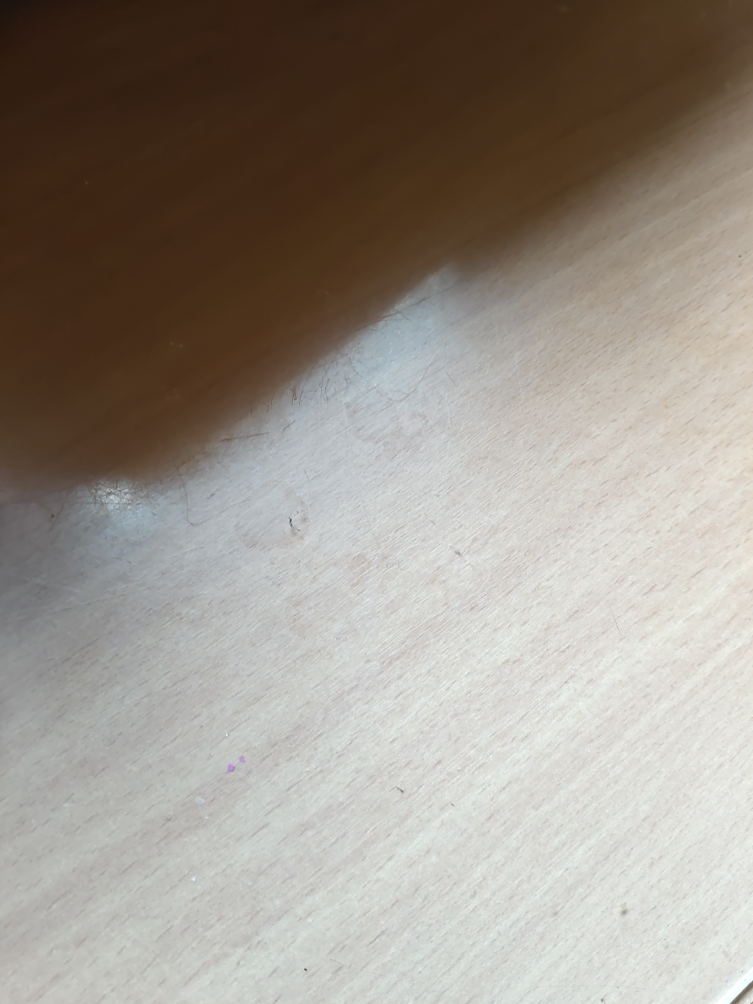 The image shows a close-up, angled view of a light-colored wooden surface, possibly a table or desk. The surface exhibits a wood grain pattern and appears slightly worn in places. A darker, shadowed area occupies the upper left portion of the image, suggesting an overhead light source or object casting a shadow. There are a few small specks, some appearing as dust or debris, and two tiny purple particles are visible near the center-right of the frame. There are no people or discernible actions depicted in the image. The location of Платошино, Russia is not determinable from the visual information provided.