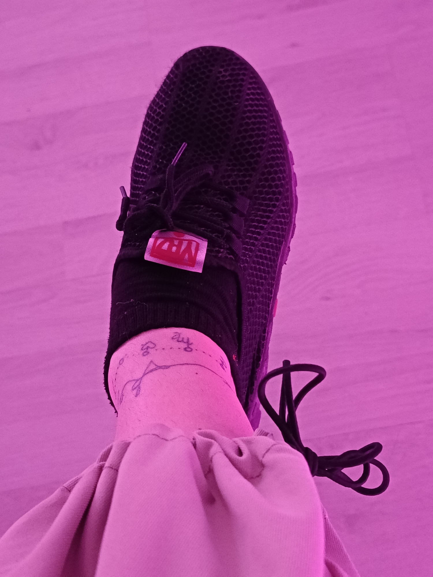 A top-down shot shows a person's foot wearing a black sneaker with black laces. The shoe has a red tag with white lettering on the tongue. The person is wearing a black sock. An ankle tattoo is visible above the sock. The person is also wearing tan pants, which are gathered around the ankle. The background is a light-colored wooden floor. The image is lit with pinkish light. The location is in the city of Unknown, Unknown.