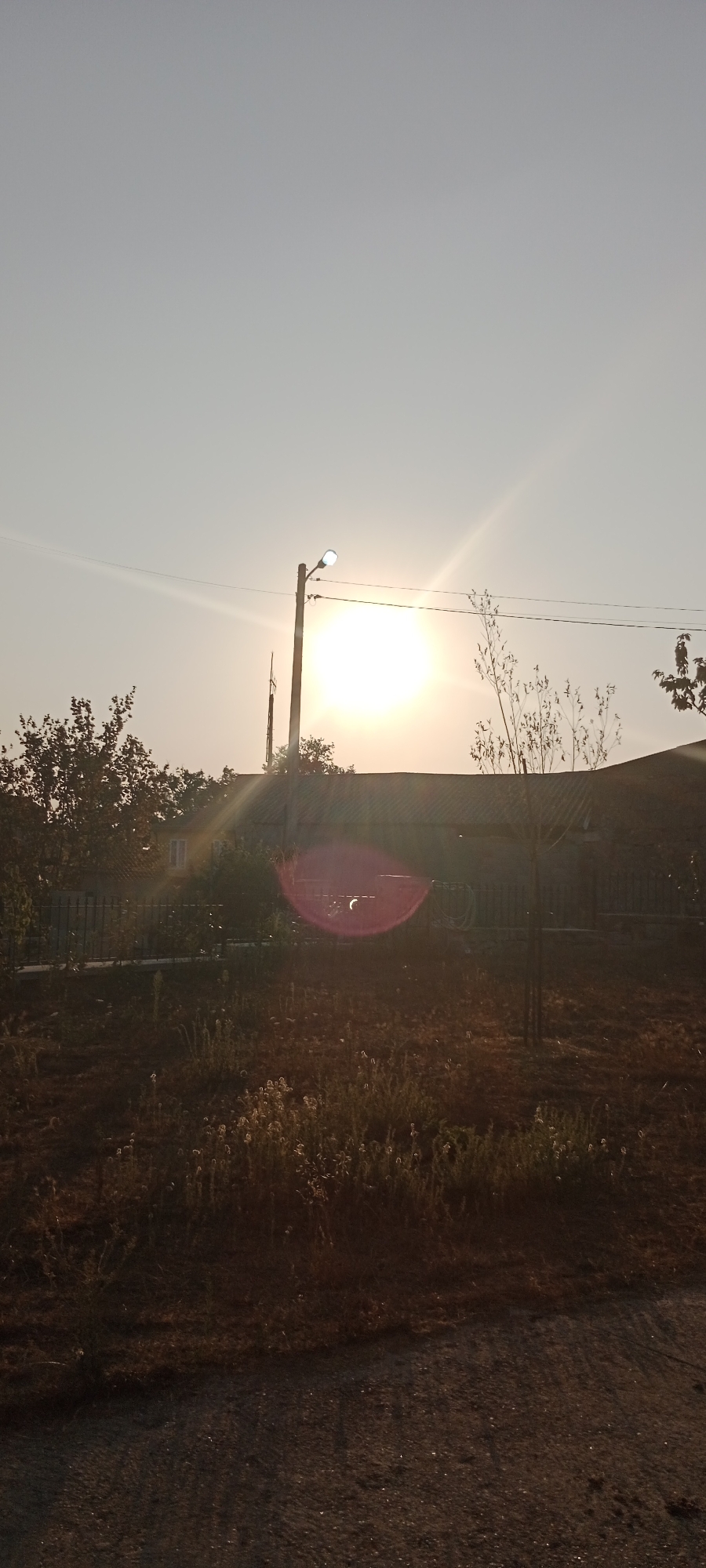 The image depicts an outdoor scene, likely in a rural or suburban setting, bathed in the warm light of a setting or rising sun. The sky is a pale, hazy blue, with the sun prominently featured as a bright, circular orb emitting strong rays. A street lamp, also illuminated, stands to the left of the sun, its light contributing to the overall brightness.
In the middle ground, a dark-roofed building, possibly a house or shed, is visible. A metal fence runs along the front of the property. To the left of the building, a leafy tree is present, its branches silhouetted against the sky. A smaller, bare-branched tree stands to the right of the building.
The foreground is a mix of dry grass, weeds, and what appears to be dirt or gravel. The ground is uneven and appears somewhat unkempt. The overall impression is one of a tranquil, possibly late summer or early autumn day. There are no people visible in the image. The provided location "Adai, Spain" does not appear to be in Adai, Spain, as there is no city named Adai in Spain.