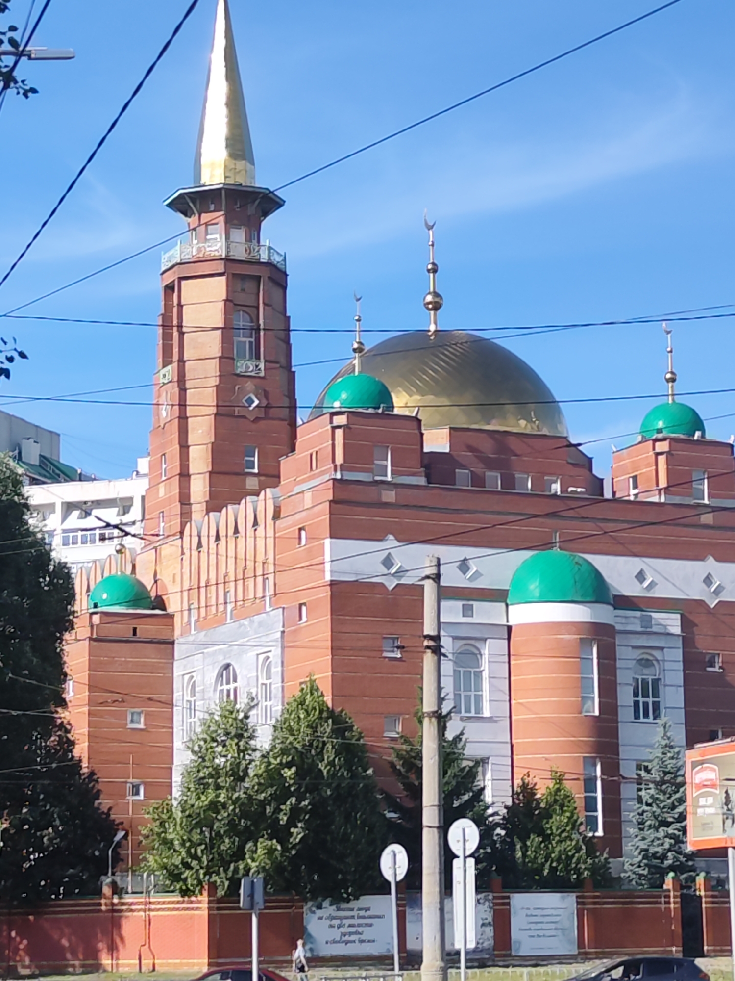 The image captures a view of the Samara Mosque, a prominent landmark in Samara, Russia. The mosque features a tall, slender minaret topped with a golden spire. Several domes, some green and one large golden one, adorn the building. The architecture is characterized by red brickwork interspersed with white decorative elements. Power lines are visible crisscrossing the sky in the foreground, partially obscuring the view.
The scene appears to be set during the daytime under a clear blue sky, suggesting fair weather. The sun casts shadows, indicating it is likely mid-morning or afternoon. Trees with lush green foliage surround the mosque, adding a natural element to the urban landscape. A street and a low wall are also visible in the lower part of the image. There are a few people visible in the distance, appearing to walk along the street. Some signage with text is present, but it is too small and blurry to be read. The overall mood of the image is serene and showcases the beauty of the mosque against a bright sky.