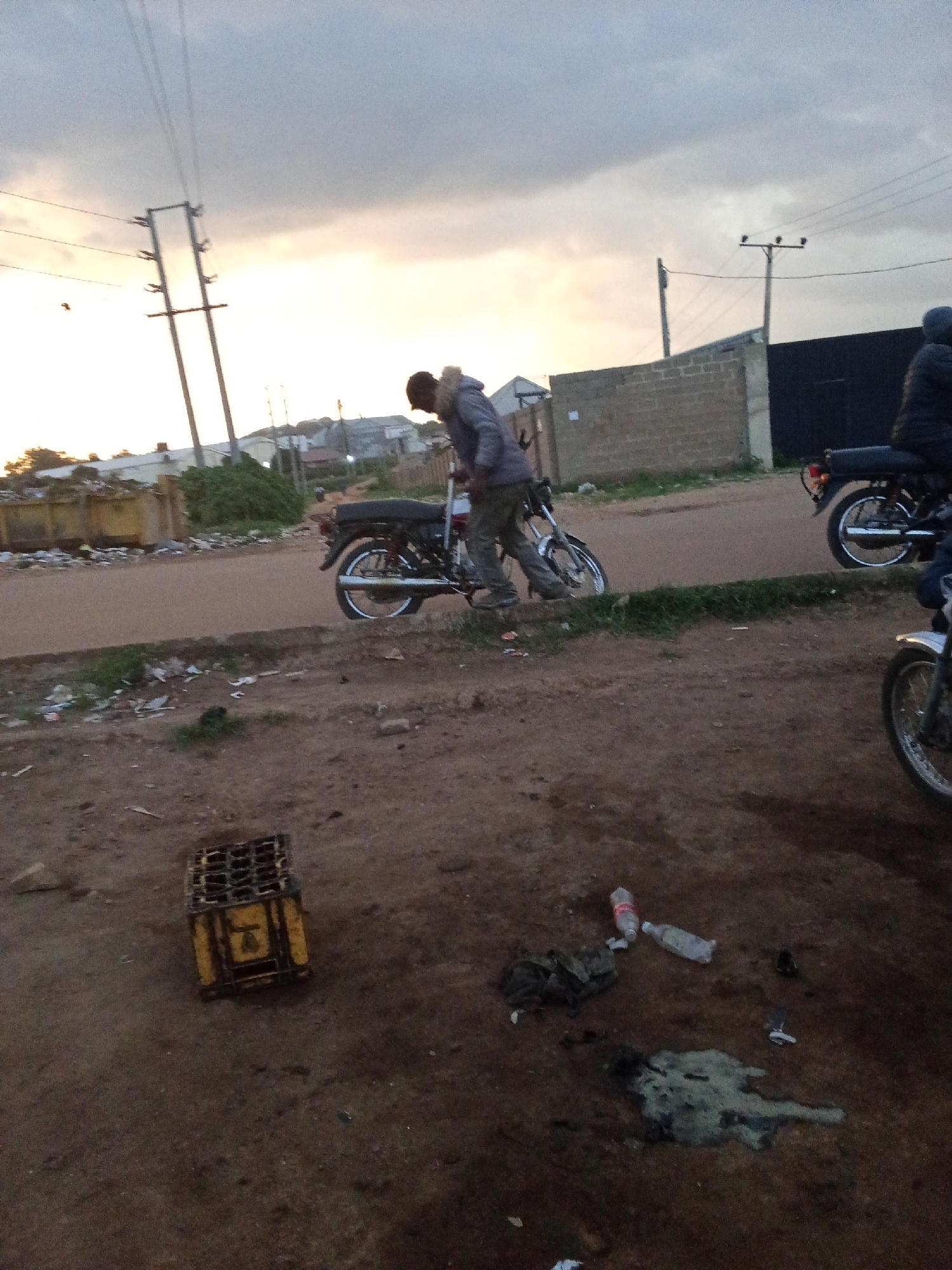 In Barakin Lerawi, Nigeria, a young man in a hooded jacket with a fur-lined collar is standing next to a motorcycle on a dirt road. The sky is overcast with hints of sunset. A brick wall and a dark fence are visible in the background, as are utility poles with overhead wires. To the left, a large yellow dumpster overflowing with trash sits near a paved road. The foreground is a dirt ground littered with debris, including a yellow crate, plastic bottles, and what appears to be spilled liquid. Another motorcycle and a person on it are partially visible in the lower right corner.