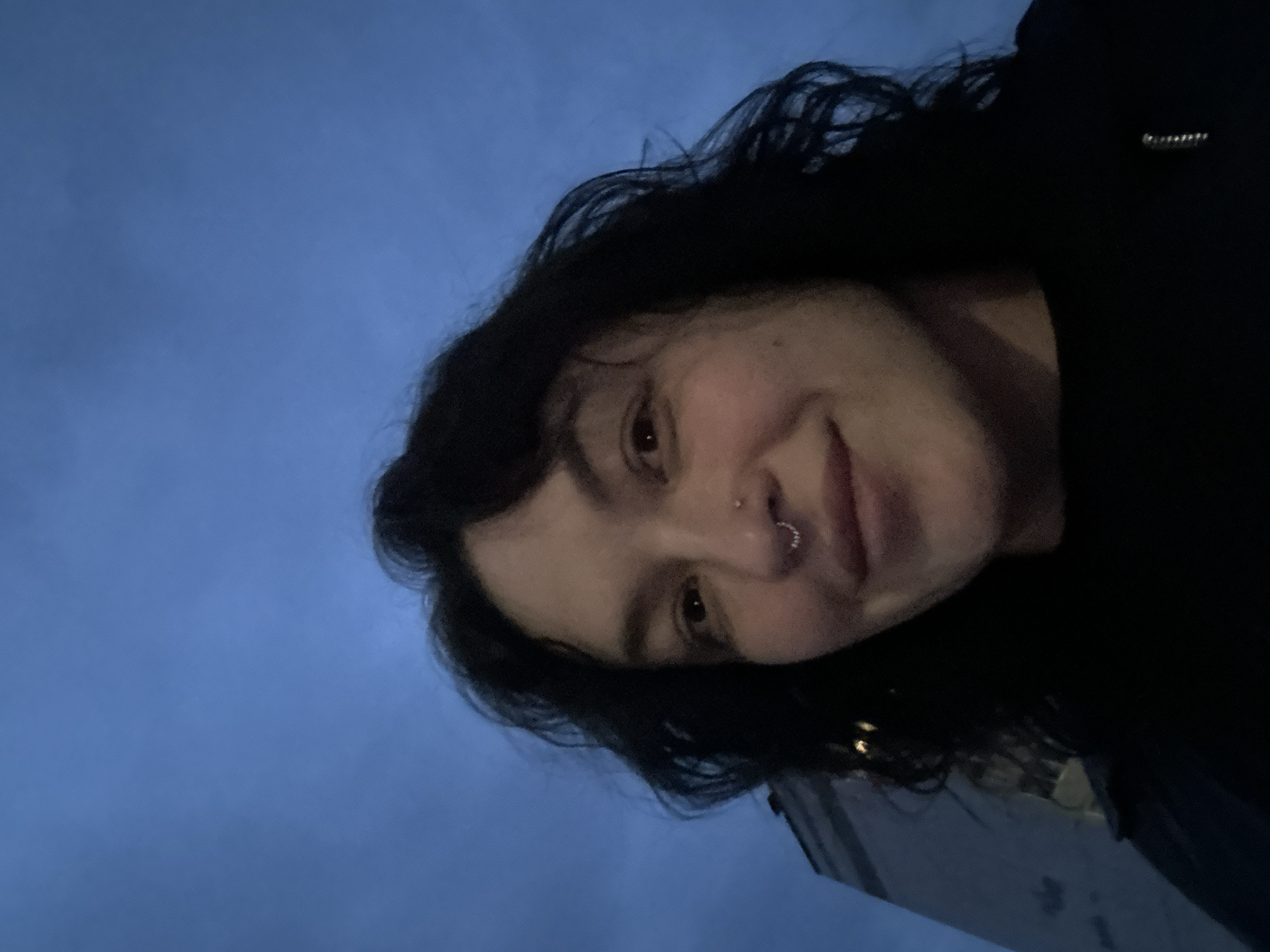 A selfie taken at an angle features a person with dark hair and a nose ring, against a background of a blue-gray sky and the corner of a light-colored building. The location is inferred as the city of Chaikovsky, Russia.