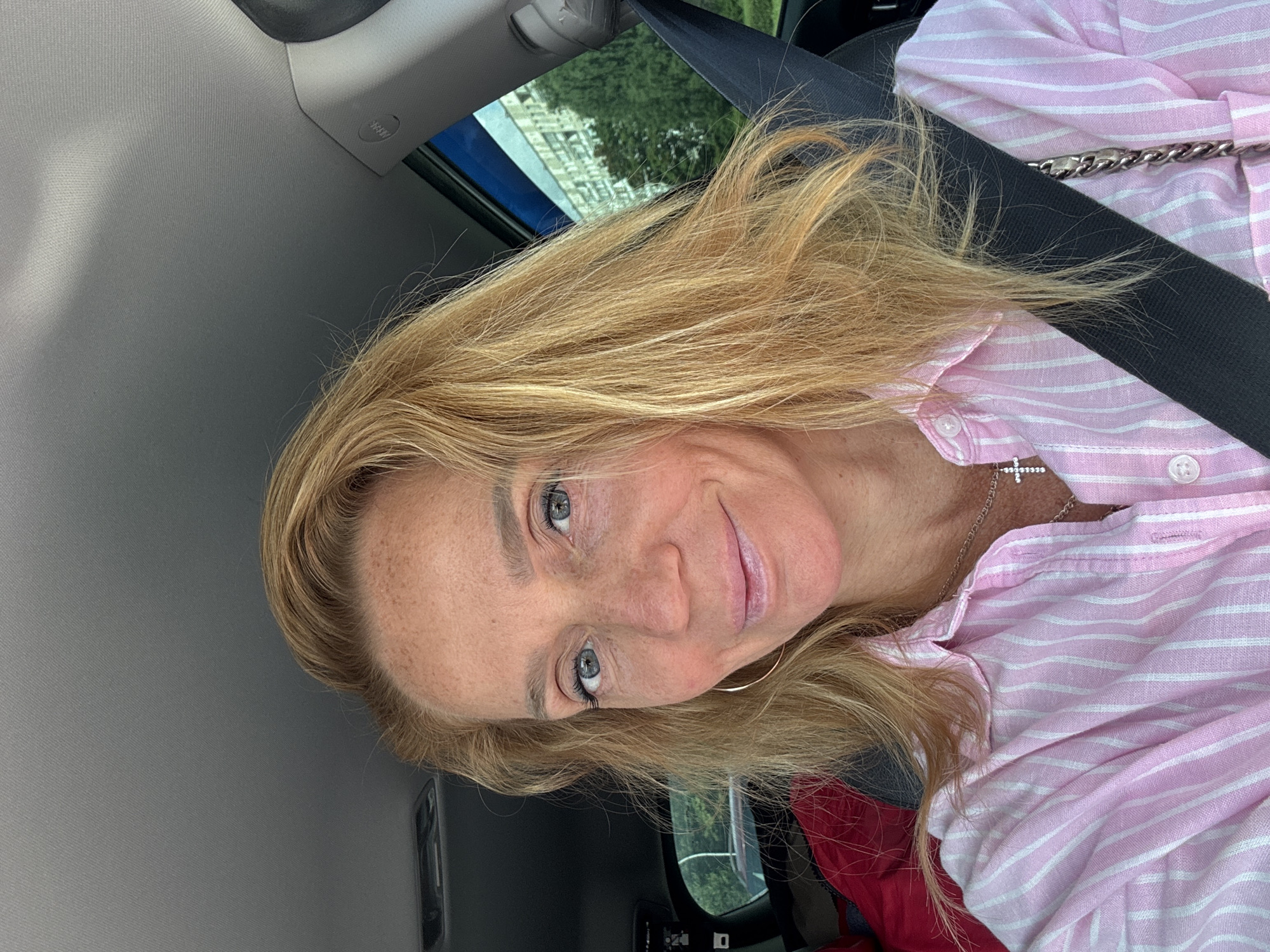 In Sinyavino, Russia, a blonde woman with light blue eyes and freckles is pictured in a car. She is wearing a pink and white striped shirt and a silver necklace with a cross pendant. Her hair is styled loosely around her face. A seatbelt is visible across her chest. Outside the car window, a building and trees can be seen.