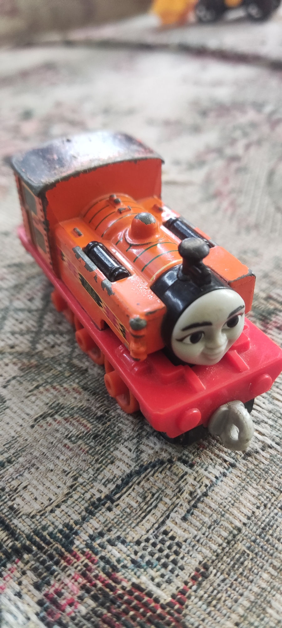A toy train, painted orange with black and red accents, is placed on a patterned cloth surface. The train features a face with large eyes and a smiling mouth. The train's paint is chipped in several areas, revealing a metallic silver underneath. The cloth surface has a woven texture with a pattern of black, beige, and red threads. The lighting suggests an indoor setting, possibly a home. There is no discernible connection to the city of Gebang, Indonesia.