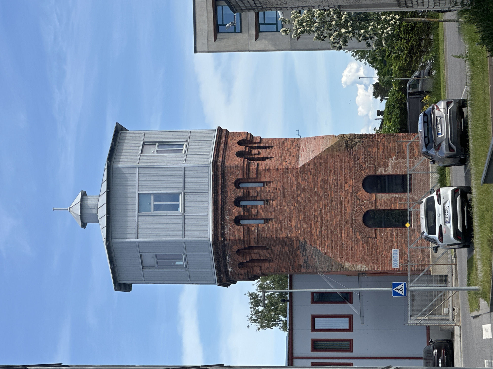 The image shows a historic red brick water tower with a modern grey upper section and conical roof. The tower is situated in a built-up area, likely an industrial or commercial district, in Tallinn, Estonia. To the left of the water tower is a grey building with tall, narrow windows. A fence separates the water tower and the grey building from a street where two cars are parked. The car closest to the viewer is a white SUV, and to its right is a silver hatchback. In the background, behind the water tower and fence, is a dense line of green trees. Further to the right, a modern concrete building with dark windows is visible, and a flowering tree with white blossoms is in the foreground. The sky is blue with scattered white clouds. A sign on the fence reads "KONTOR", which is Estonian for "Office." A blue and white pedestrian crossing sign is also visible on a pole.