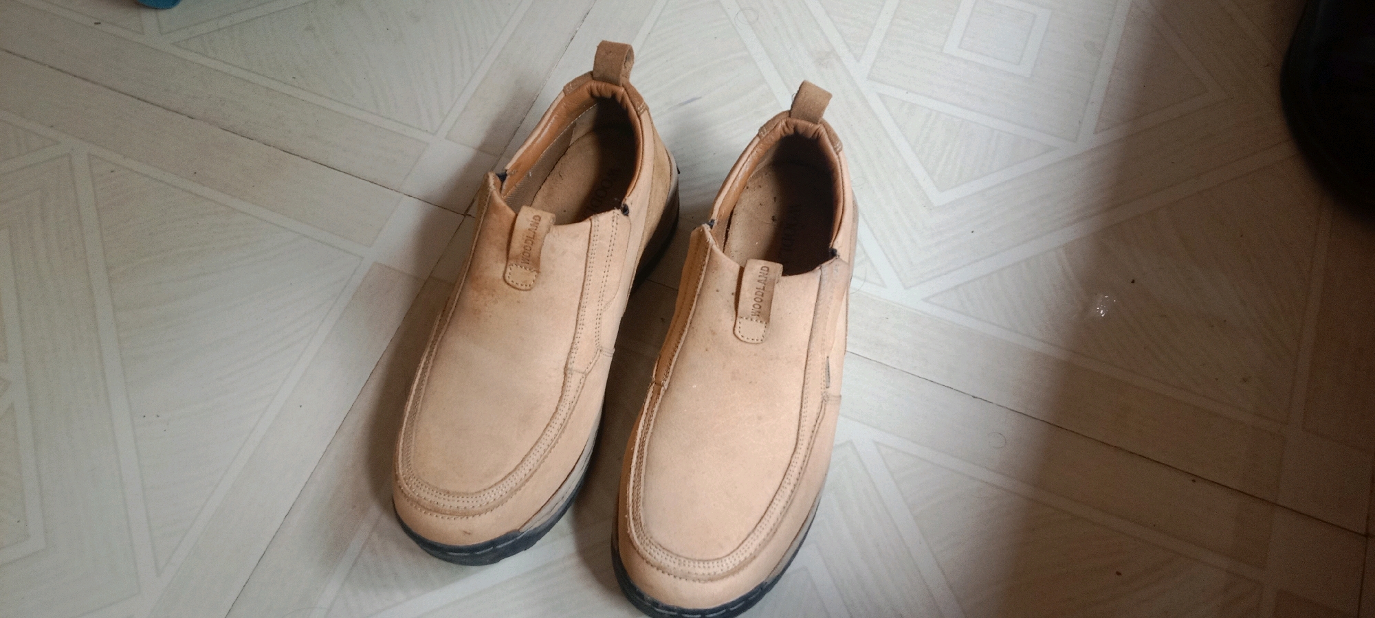 A pair of tan, casual slip-on shoes with a textured sole are displayed from a top-down perspective on a light-colored tiled floor. The shoes are positioned side-by-side, with the left shoe slightly overlapping the right. The brand name "WOODLAND" is visible on a small tag on the upper of each shoe. The floor appears to be indoors, with a geometric pattern formed by the tiles. The image is taken in Varkala, India.