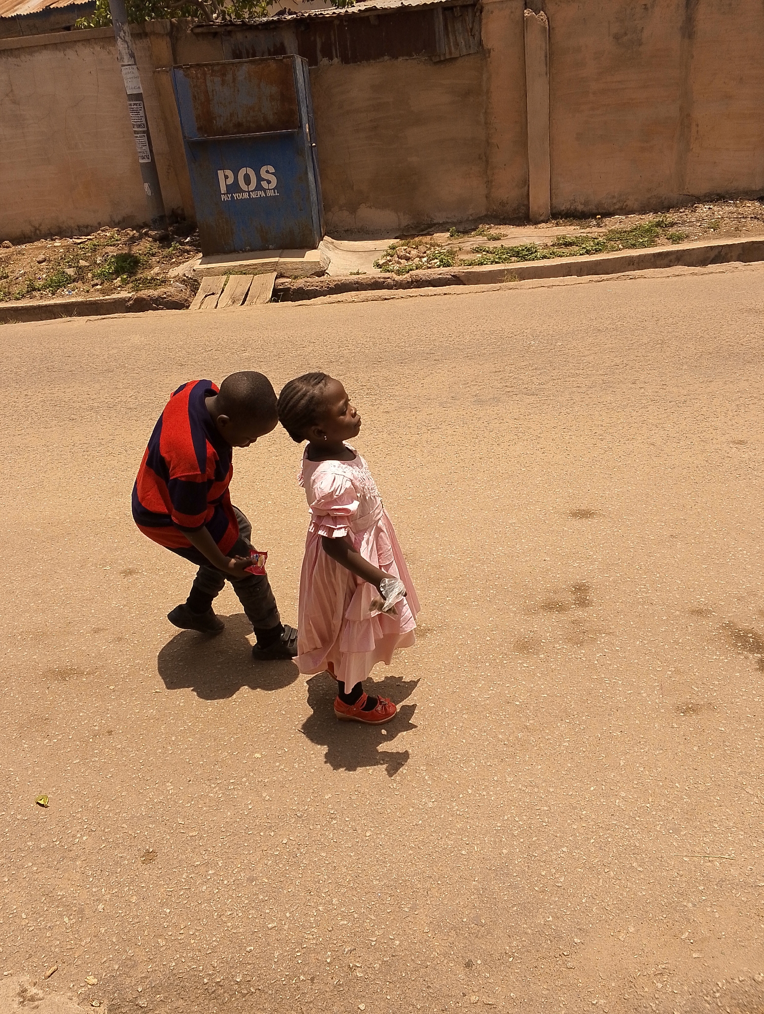 Two children stand on a paved road in Jos, Nigeria. The boy, dressed in a red and blue striped shirt, dark jeans, and black shoes, is bending over and holding a small package. The girl, wearing a pink dress and red sandals, is looking up and to the right. Behind them, a light brown concrete wall is topped with a corrugated metal roof. A blue metal utility box with the inscription "POS PAY YOUR NEPA BILL" sits next to the wall. A utility pole is visible to the left.