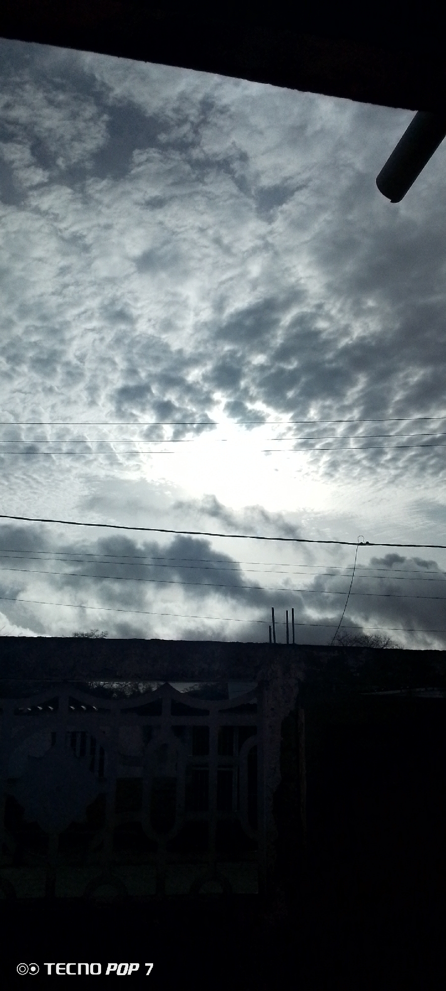 The image captures an outdoor scene in the city of Altagracia, Venezuela, under a heavily overcast sky, with a strong light source, likely the sun, obscured behind dense clouds in the upper central-right portion, causing significant backlighting. The sky is dominated by various shades of grey stratocumulus or altocumulus clouds, with the light source creating a bright, almost blown-out area. Multiple dark utility lines traverse horizontally across the mid-section of the frame. Below these lines, a dark, silhouetted distant landscape or low hills are visible, topped with sparse, dark vegetation. In the immediate foreground, a dark, solid horizontal structure, possibly a wall or roofline, is present, from which three thin vertical posts protrude on the right. Below this, a decorative, heavily shadowed fence or railing with geometric patterns is partially visible. A dark, diagonal cylindrical pipe extends downwards from the upper-right corner, and a dark overhang frames the very top. The scene depicts an urban or semi-urban environment, characterized by man-made structures and utility infrastructure, rendered largely in a desaturated palette due to the backlighting. No individuals are visible.