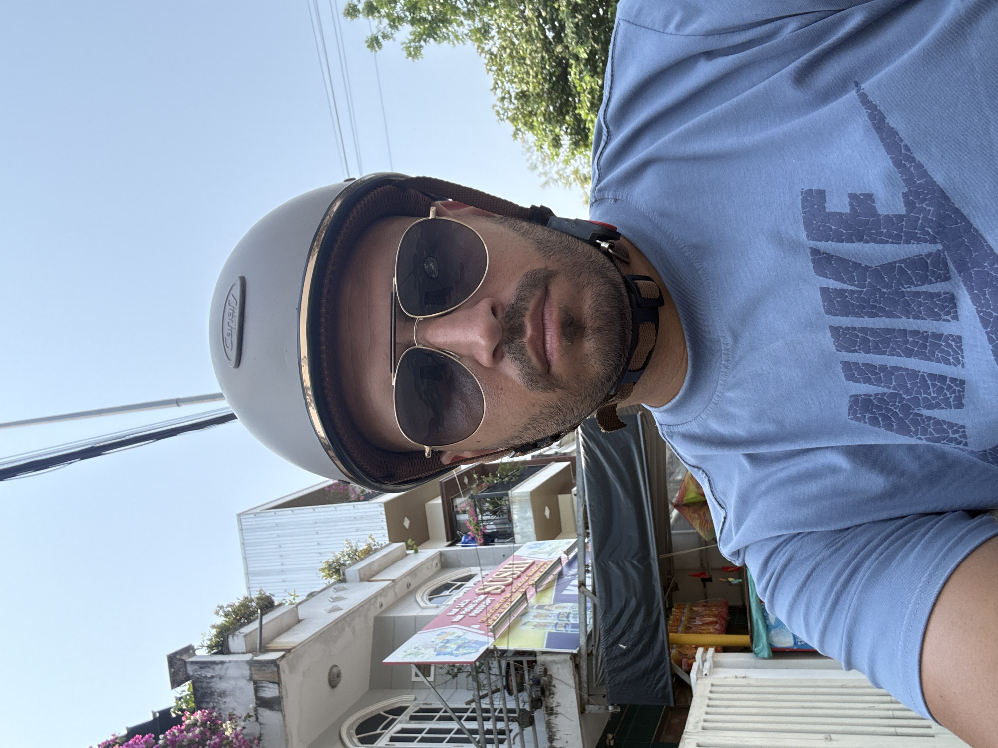 A man wearing a light blue t-shirt, a grey helmet, and sunglasses is taking a selfie. The t-shirt has a faded graphic of a Nike logo. The helmet's strap is visible. The background shows a street scene in what appears to be Đà Nẵng, Vietnam. A building with white walls, rounded windows, and a sign that reads "SUSHI" is visible. Bougainvillea flowers are growing around the building's exterior. Overhead power lines are also in view. The lighting suggests it is daytime.