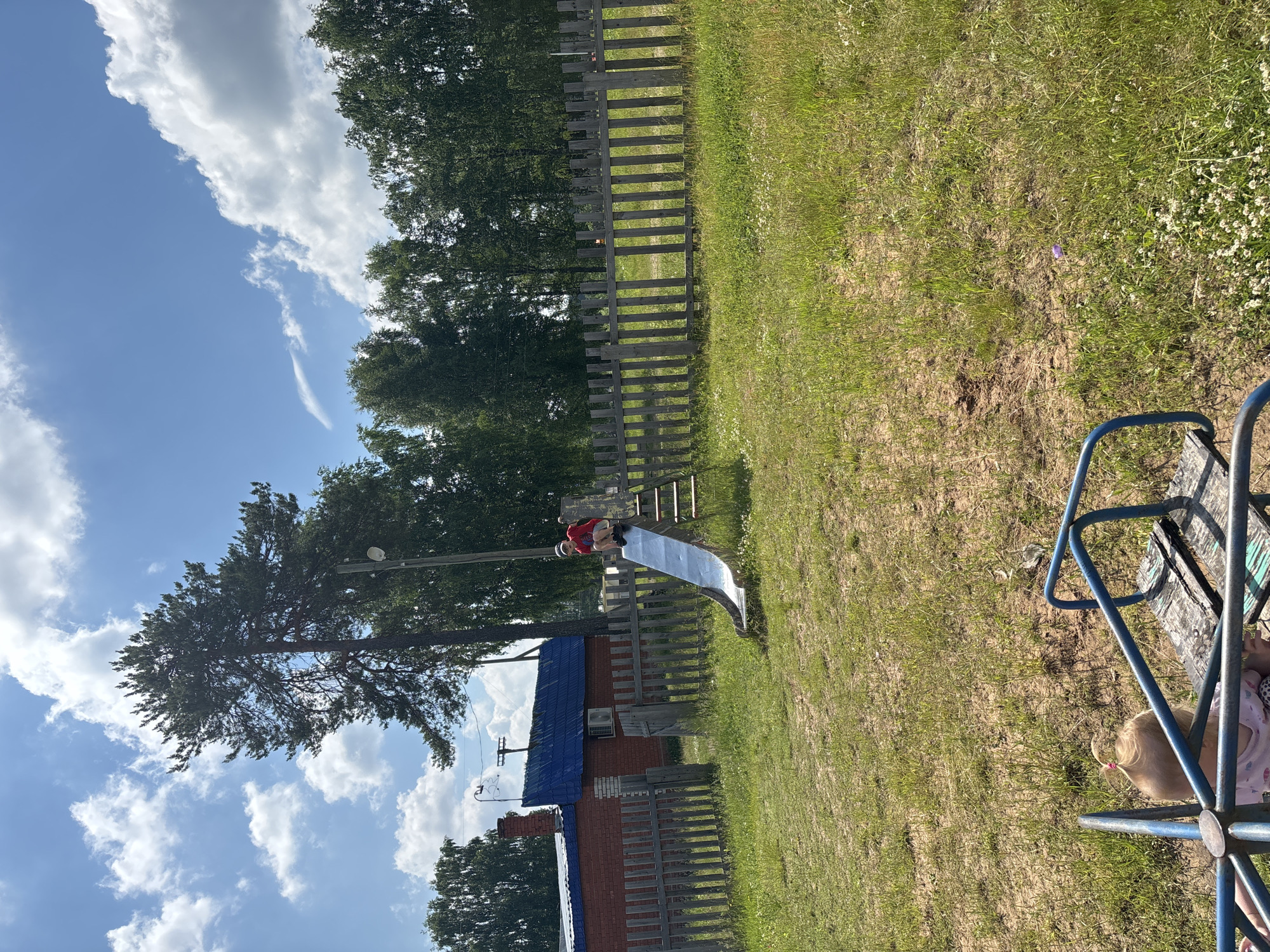 A child is sliding down a metal slide in a grassy yard. The child is wearing a red shirt and shorts, and a hat. The slide is on a playground structure with wooden steps. In the background, there is a wooden fence, a brick building with a blue roof, and trees. The sky is blue with white clouds. There is also a merry-go-round in the foreground, with a baby on it. The scene appears to be in a rural area, possibly a backyard or a small park. Based on the provided context, this scene is likely located in the city of Kurgan, Russia.