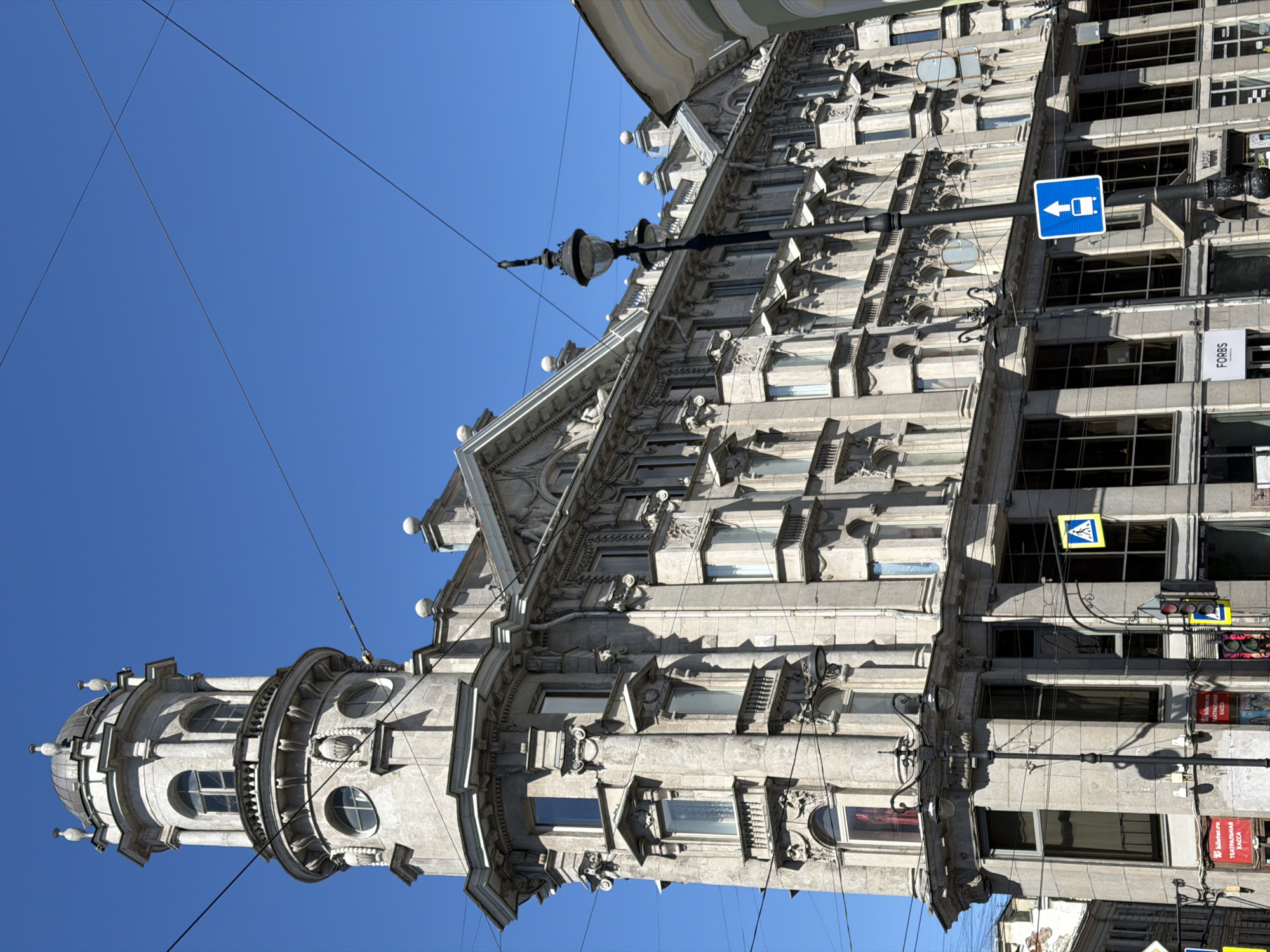 This image captures the Eliseev Merchants' House (Eliseyev Shop) located at the intersection of Nevsky Prospekt, 56, and Malaya Sadovaya Street, 10, in Saint Petersburg, Russia. The prominent, multi-story building is constructed of light grey stone and showcases intricate Art Nouveau and Neoclassical architectural details. A distinctive circular tower, featuring arched and round windows, decorative balustrades, and a domed top adorned with finials, crowns the building's corner. The facade is richly ornamented with columns, pilasters, balconies with railings, and various relief carvings.
The scene is illuminated by bright daylight under a clear blue sky. In the foreground, an ornate black street light pole with multiple luminaires stands, from which numerous black overhead utility wires extend and crisscross the upper portion of the frame. On the lower section of the building, large rectangular windows are visible, some displaying reflections. Commercial signs for "FORBS," "Билетер.ру" (Bileter.ru), and "Театральная КАССА" (Theater Box Office) are affixed to the ground floor. Traffic signs, including a blue square with a white triangle and walking person (pedestrian crossing), and a blue square with a white upward-pointing bus icon (bus stop), are mounted on poles. A traffic light is partially visible on the left. No people are present in the image.