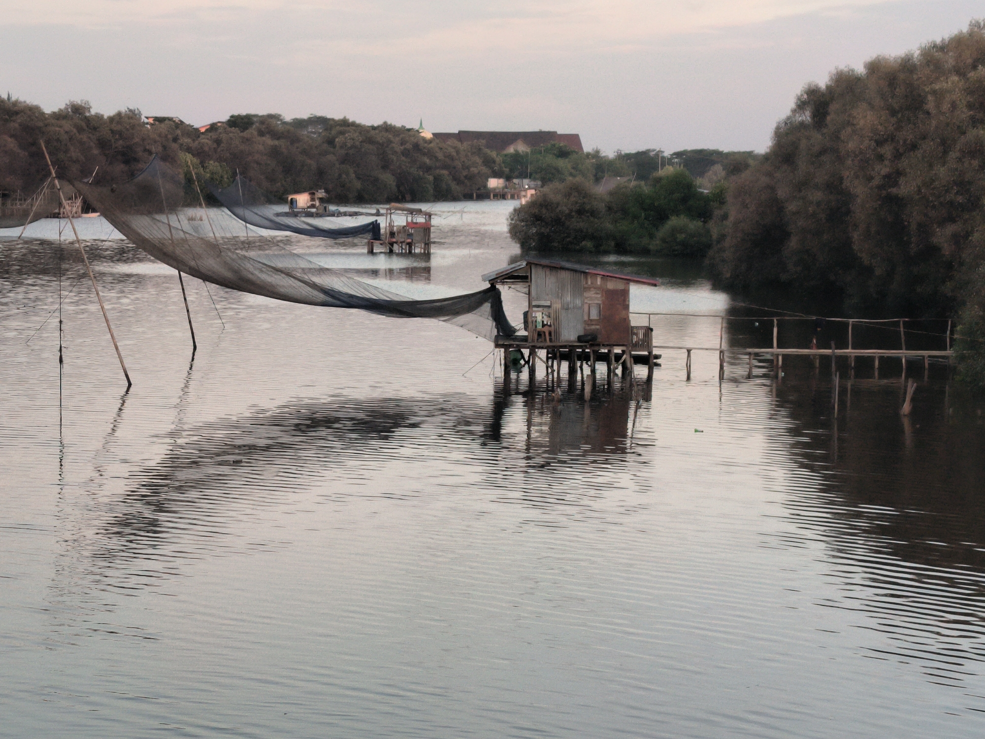 This image depicts a tranquil scene on a body of water, possibly a river or lake, in Lhokseumawe, Indonesia. Several wooden structures on stilts are visible, each topped with a small shack or hut. A large fishing net hangs suspended above the water, held up by tall poles. The surface of the water is rippled, reflecting the structures and nets, creating a blurred reflection. Lush greenery and trees line the banks of the water, adding depth to the scene. The sky is overcast, providing soft and diffused lighting to the landscape. The overall atmosphere suggests a quiet, working environment of a fishing community. No people are directly visible, but the structures suggest human presence and activity.