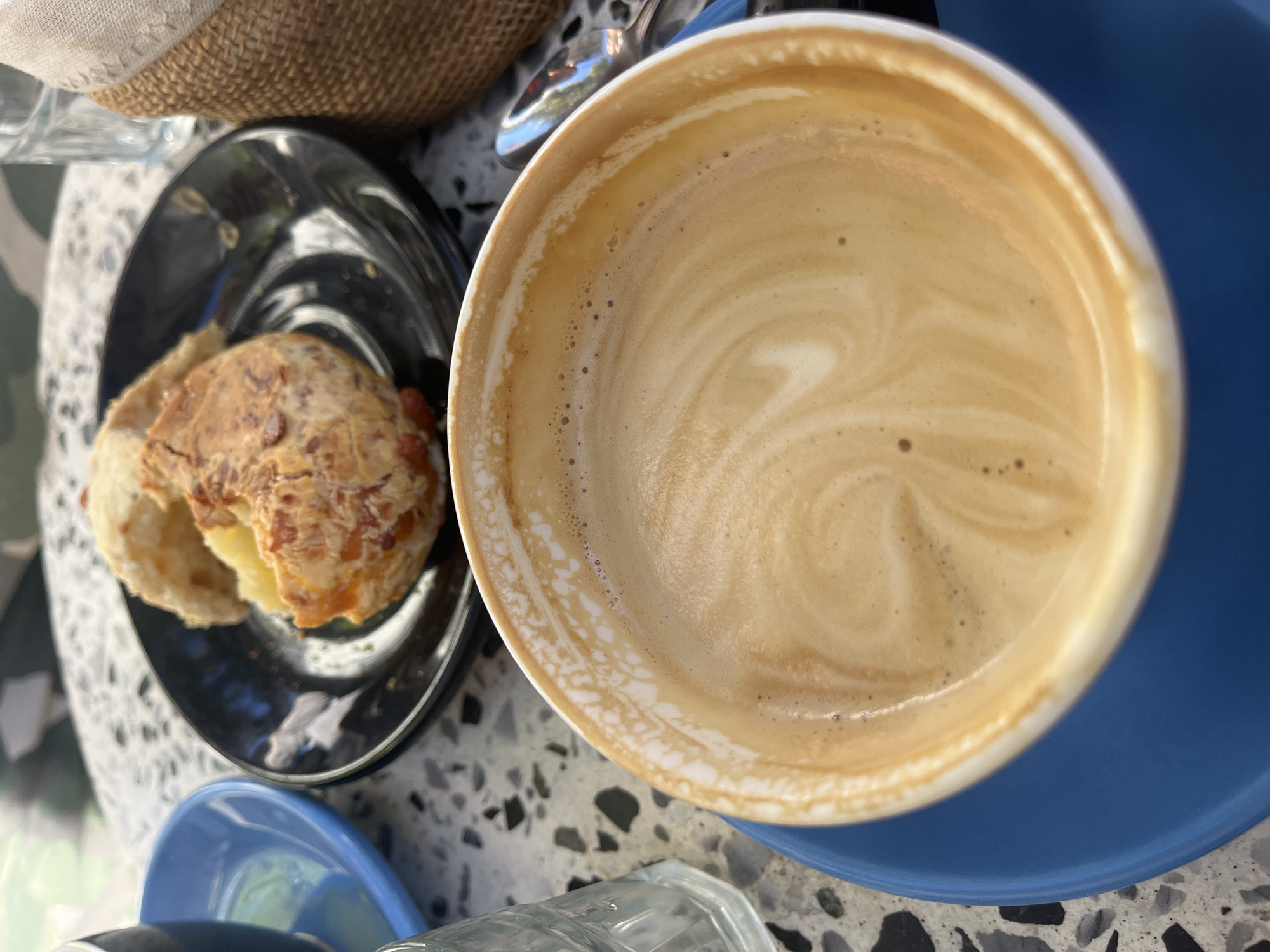 A high-angle shot reveals a cup of coffee on a blue saucer, alongside a black plate with a partially eaten pastry, all resting on a terrazzo table in Olivos, Argentina. The coffee cup is white with a tan interior, showing swirling latte art and small bubbles. The pastry on the black plate appears to have cheese baked on top and a white filling. The terrazzo table has a mix of black, white, and gray fragments. A woven basket and a glass are partially visible in the background.