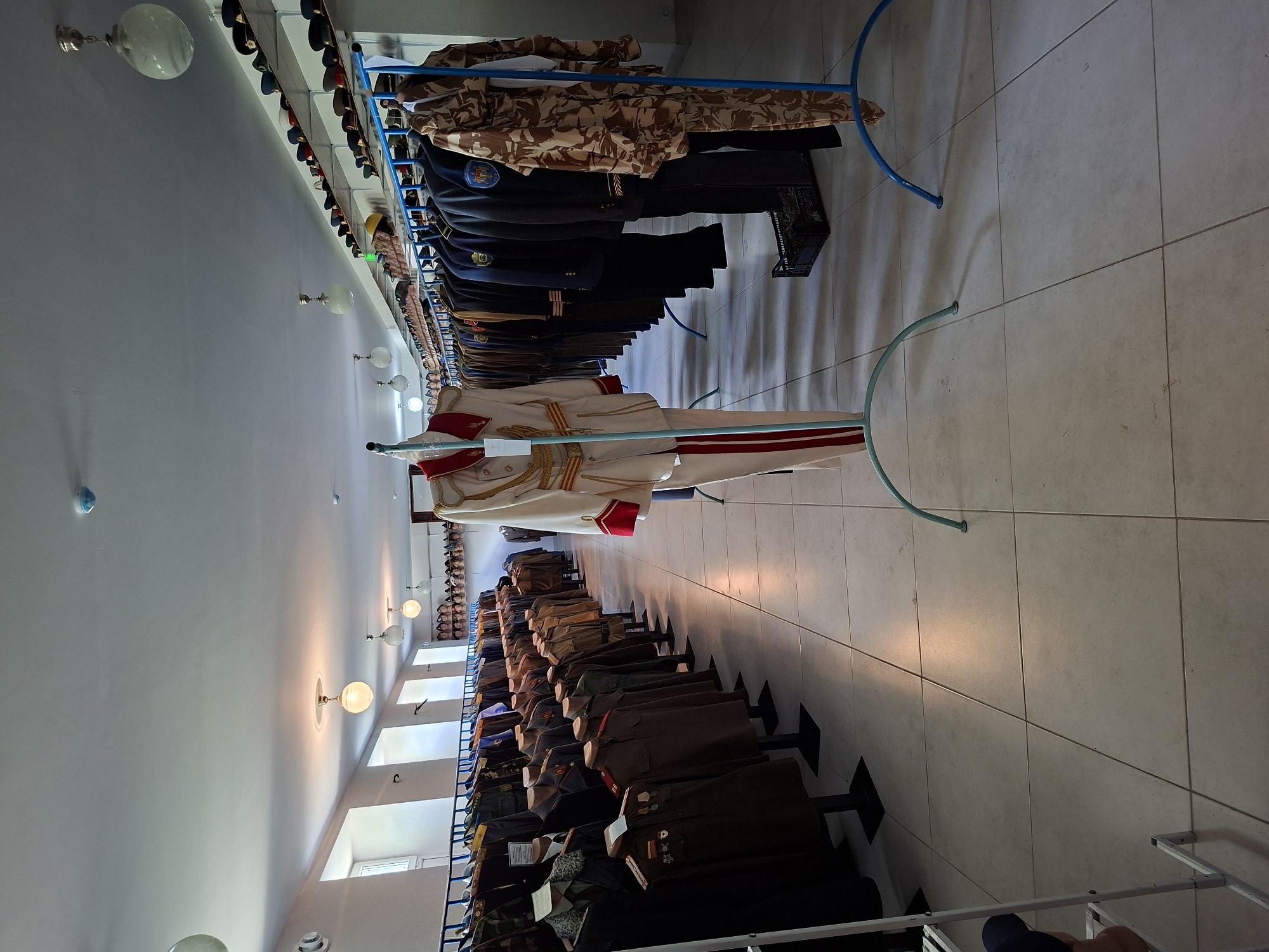 The image depicts the interior of a museum exhibition, likely the "House of Culture" in Ialoveni, Moldova, showcasing a collection of historical uniforms and military attire. Rows of mannequins are displayed, dressed in various formal military uniforms, some with medals and insignia. These are arranged on clothing racks, with a prominent white and red ceremonial uniform on a display stand in the foreground. Shelves above the racks are filled with a collection of hats, many appearing to be military caps. The room has a tiled floor, white walls, and is illuminated by overhead light fixtures and natural light from windows. The overall impression is of an organized display of military history.