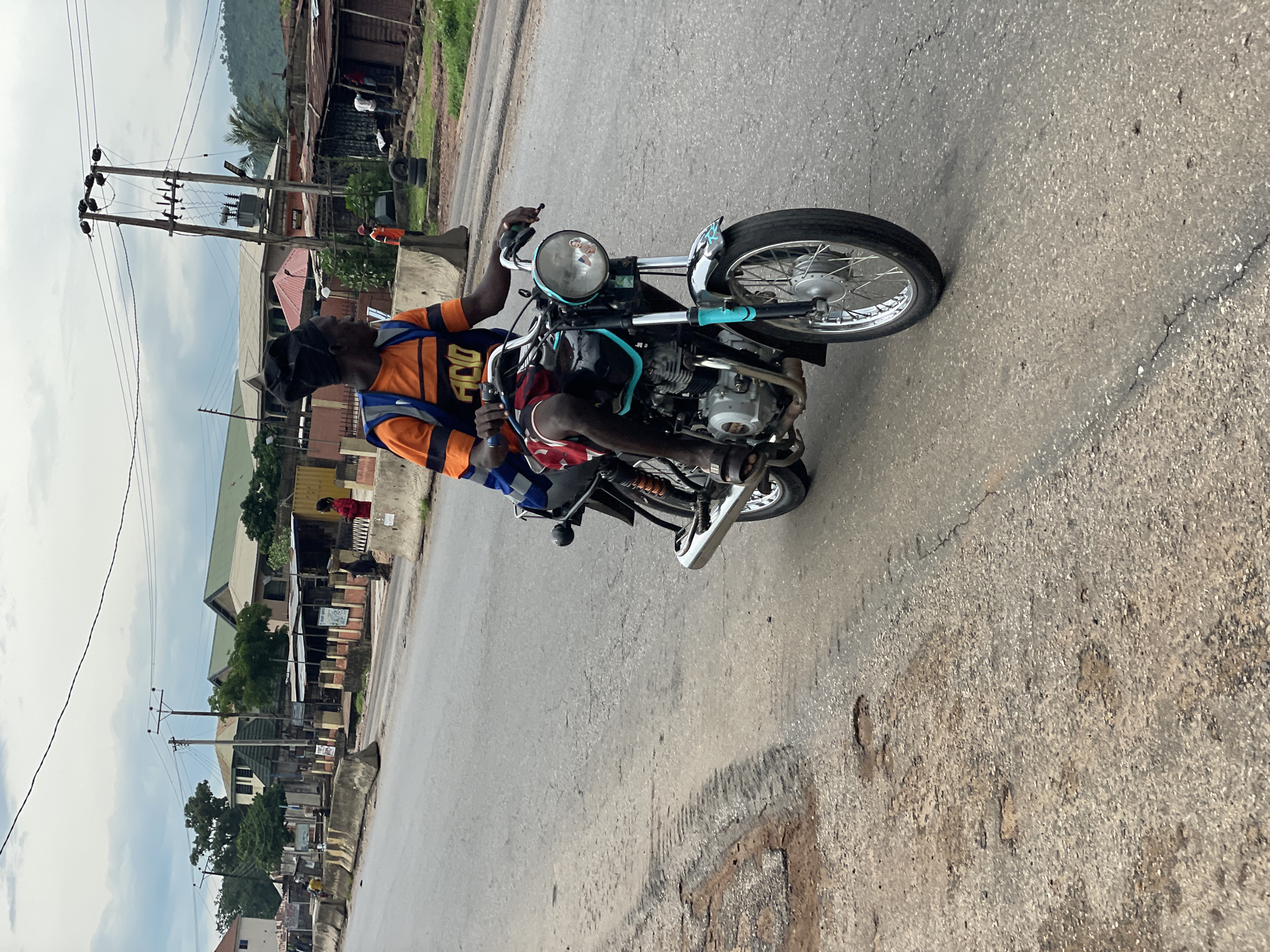 A man is riding a motorcycle down a paved road. He is wearing an orange and blue shirt with "FICO 400" written on it, and a black head covering. The motorcycle has a blue trim on the handlebars and a reflection of a person on the headlight. The road is lined with buildings and power lines, indicating an urban or semi-urban setting. The sky is overcast, suggesting it might be a cloudy day. In the background, there are trees and a hill. The scene appears to be in Lokoja, Nigeria, as per the location context provided.