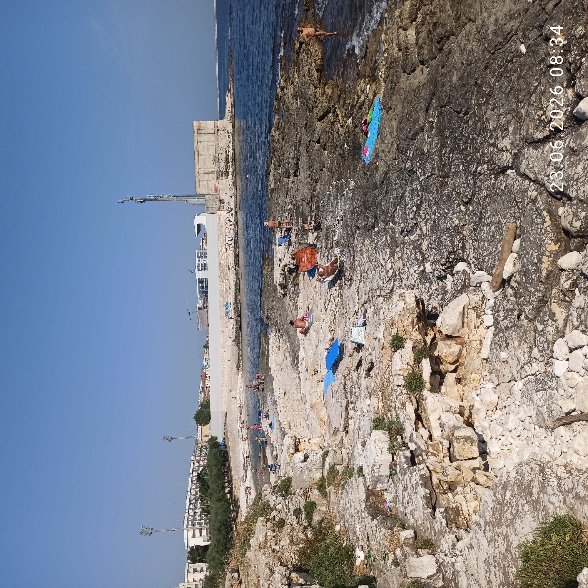 The image captures a rocky coastline near Molfetta, Italy, with several individuals enjoying the sunny weather. In the foreground, the rough, light-colored rocks are dotted with sparse vegetation. Several people are visible on the rocks, some sunbathing on towels and loungers, while others are near the water's edge.
Further back, a small sandy beach is visible, with more people swimming and relaxing. In the background, a long, modern-looking building or structure runs along the coastline, with a tower equipped with antennas. Trees and residential buildings are situated behind this structure, suggesting the proximity of an urban area. The sea is calm and blue, reflecting the clear, cloudless sky. The timestamp "23.06.2026 08:34" is superimposed on the lower right corner of the image.