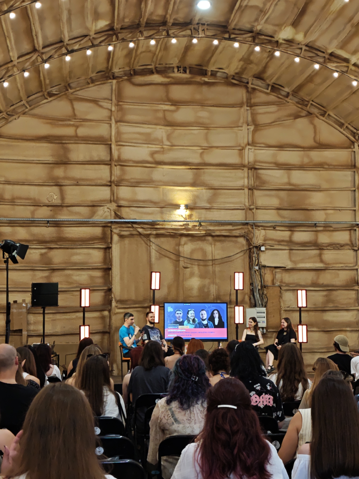 The image depicts an indoor event, possibly a conference or a presentation, held in a large, hangar-like space. The architecture suggests a rustic, industrial aesthetic, with exposed wooden beams forming a curved ceiling lined with string lights. The walls are also paneled with wood, creating a warm, natural color palette.
In the foreground, an audience of people is seated, facing a stage area. The audience members are diverse in age and gender, with many exhibiting colorful hair. Their backs are mostly to the camera, suggesting they are engaged with the on-stage activity.
On the stage, there are several individuals. Two men are seated in chairs on the left, seemingly participating in a discussion or presentation. A large screen behind them displays images of people. To the right of the screen, two women are seated, also appearing to be involved in the event. The overall atmosphere seems attentive and engaged.
The setting's design, particularly the prominent use of wood and the spaciousness, could align with repurposed industrial buildings often found in urban areas that are being redeveloped for cultural or event purposes. Without specific visual cues that definitively point to Lviv, Ukraine, it's impossible to confirm the location. However, such venues are characteristic of many contemporary event spaces in cities undergoing modernization.