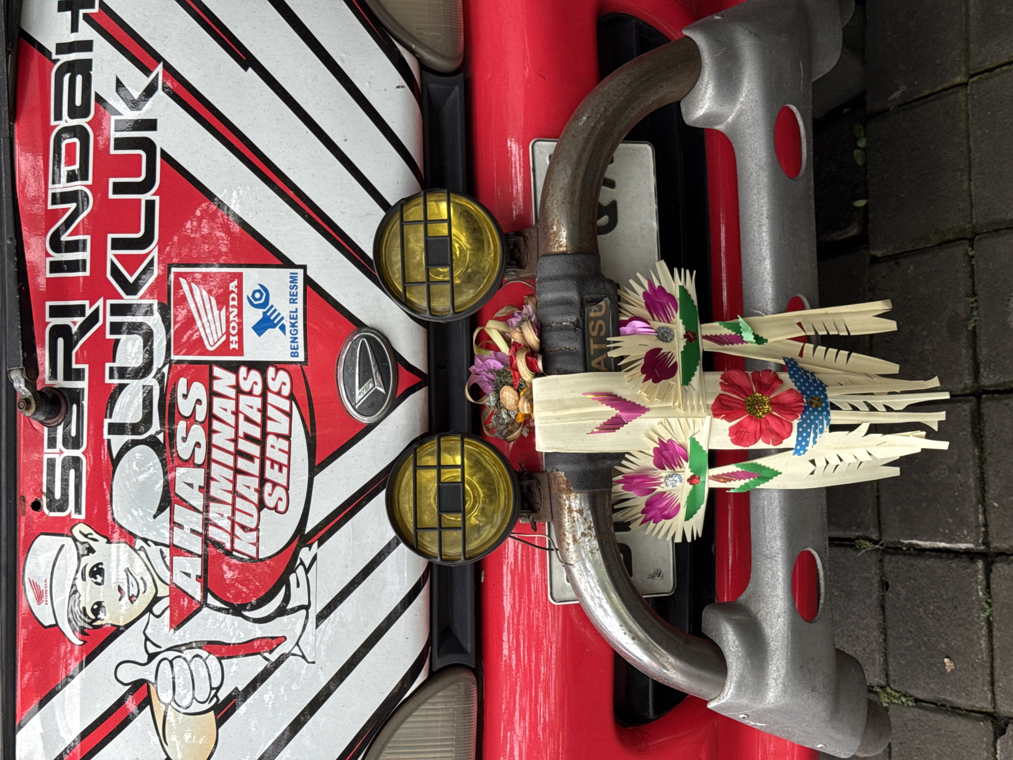 A close-up shot shows the front of a red and white vehicle, likely a motorbike or scooter, in Kuta, Indonesia. The vehicle features a Honda logo and the text "SERI INDAH KLIK" and "AHASS JAMINAN KUALITAS SERVIS." The front has two yellow, caged fog lights and a chrome Mercedes-Benz logo. A curved chrome bar adorned with traditional Balinese offerings made of woven palm leaves and decorative flowers and a small sign is attached to the front of the vehicle. The setting appears to be an outdoor, possibly urban, environment with gray brick pavers visible in the lower right of the frame.