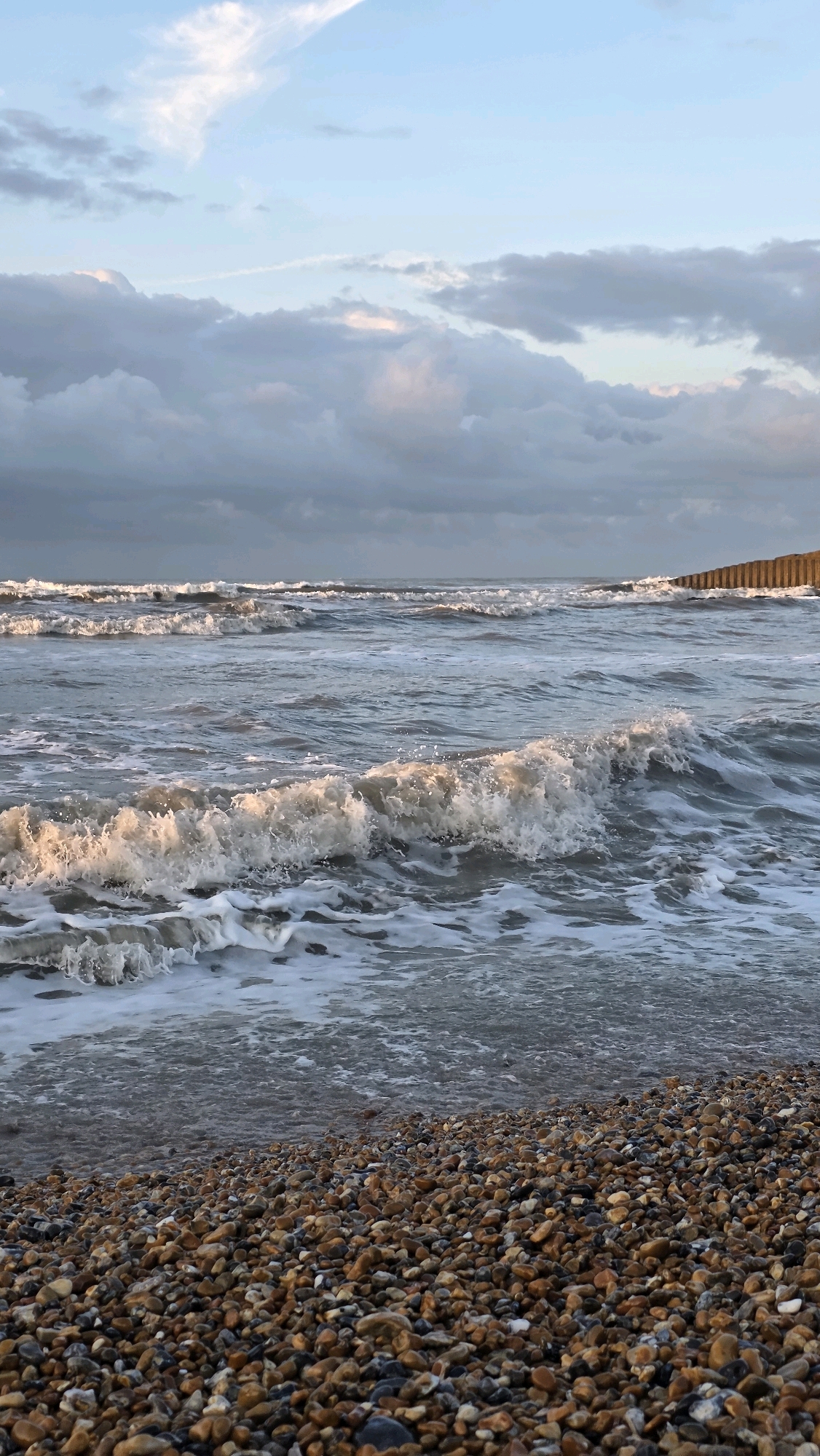 A vertical, eye-level shot captures a wave-filled beach in St Leonards, United Kingdom. The foreground consists of a densely packed, multi-colored pebble shore. Beyond the shore, the sea is characterized by choppy, grey-blue waves that break into white foam. In the distance, a wooden breakwater juts out into the sea from the right side of the frame. The sky above is a blend of pale blue and scattered grey clouds, with hints of pink and white in the clouds, suggesting either dawn or dusk. The lighting is soft, casting a diffused glow over the scene. There are no people or animals visible in the image.