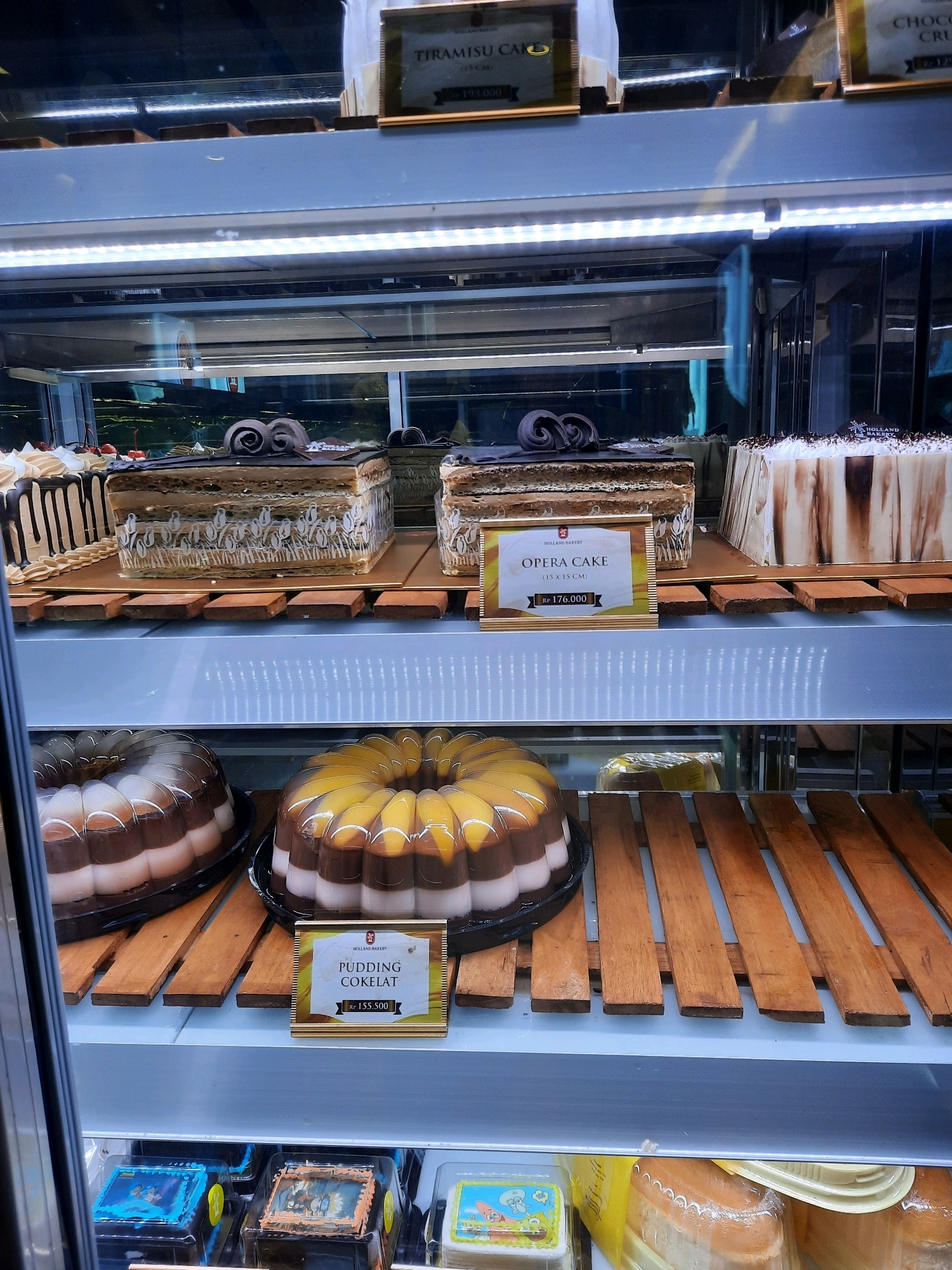 The image shows a display of cakes and puddings in a bakery case in East Jakarta, Indonesia. The bakery case is well-lit, and the products are arranged neatly on shelves.
In the top shelf, there are several cakes. One cake is labeled "TIRAMISU CAKE" and another is labeled "CHOCO CRU". To the left of these is a cake with dripping chocolate frosting and red cherries on top. To the right of the "TIRAMISU CAKE", there's another cake partially visible with chocolate swirls.
The middle shelf displays two cakes prominently. The left cake is a tall, rectangular cake with a detailed pattern on its sides and chocolate swirls on top. A sign in front of it reads "HOLLAND BAKERY OPERA CAKE (15 x 15 CM) Rp 176.000". The right cake is a large, rectangular cake with a white frosting and brown marbled design.
The lower shelf features two ring-shaped puddings. The left pudding has distinct layers of brown and white. The right pudding has layers of brown, white, and a yellowish fruit-like topping. In front of the right pudding, a sign reads "HOLLAND BAKERY PUDDING COKELAT Rp 155.500".
The bottom shelf contains several smaller square cakes in clear plastic packaging. Some of these cakes have cartoon characters or decorative images on top. There are also some larger baked goods in plastic containers visible on this shelf. The overall environment suggests a commercial bakery with a focus on attractive dessert presentation.