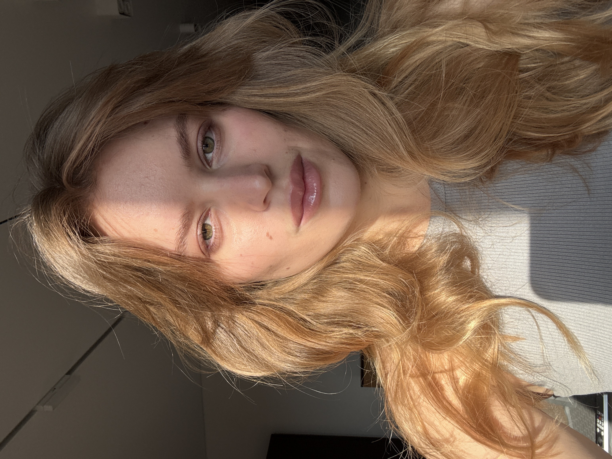 A young woman with long, wavy blonde hair is facing the camera. She has fair skin and green eyes. The lighting suggests she is near a window, possibly indoors. Her lips are glossy, and she appears to be wearing minimal makeup. She is wearing a gray top. The background is dark, and a wall is seen in the lower left corner. The image appears to be taken at an angle, resulting in a rotated perspective. The location cannot be determined in the city of Hrodna, Belarus, based solely on the visual information in the image.