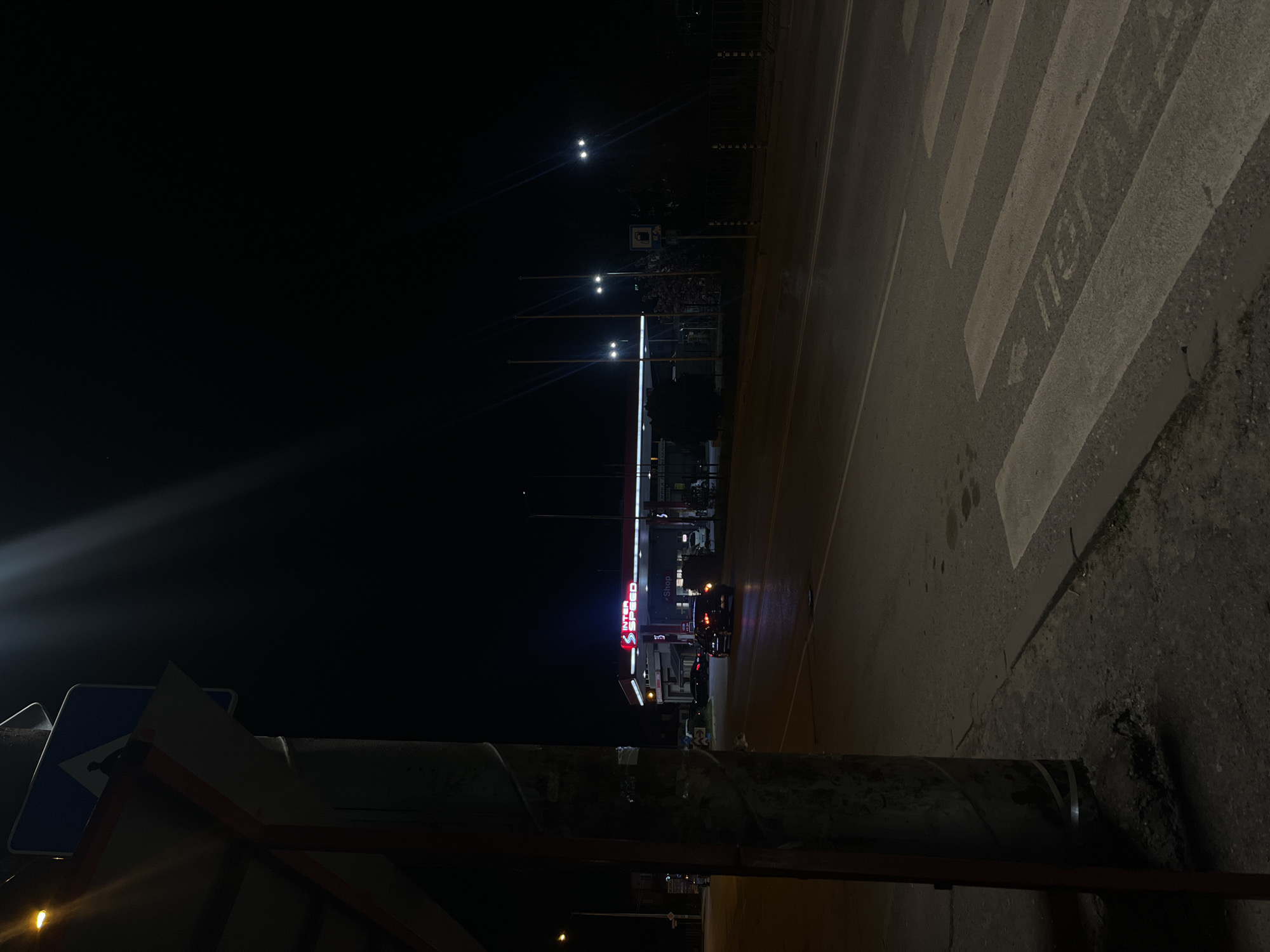 The photo was taken at night in an urban area. A brightly lit gas station is visible in the middle ground, identifiable by its red signage. In the foreground is a pedestrian crosswalk. A street sign with directional arrows is partially visible, suggesting it is an intersection or junction. A road with cars parked alongside the gas station can be seen, indicating a moderate level of activity despite the time of day. The lighting conditions create stark contrasts between the illuminated areas and the surrounding darkness. The location is believed to be Sofia, Bulgaria.