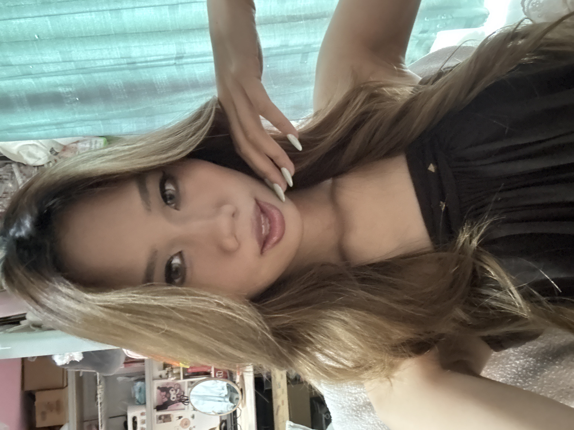 A woman with long brown hair and light skin is lying down, with her head tilted to the left. She is wearing a black dress. One hand is raised near her face. The background shows a room with curtains and shelves, and what appears to be a desk area with a mirror and various items. The location is likely an indoor setting, and the context of Nan'an District, China is extraneous.