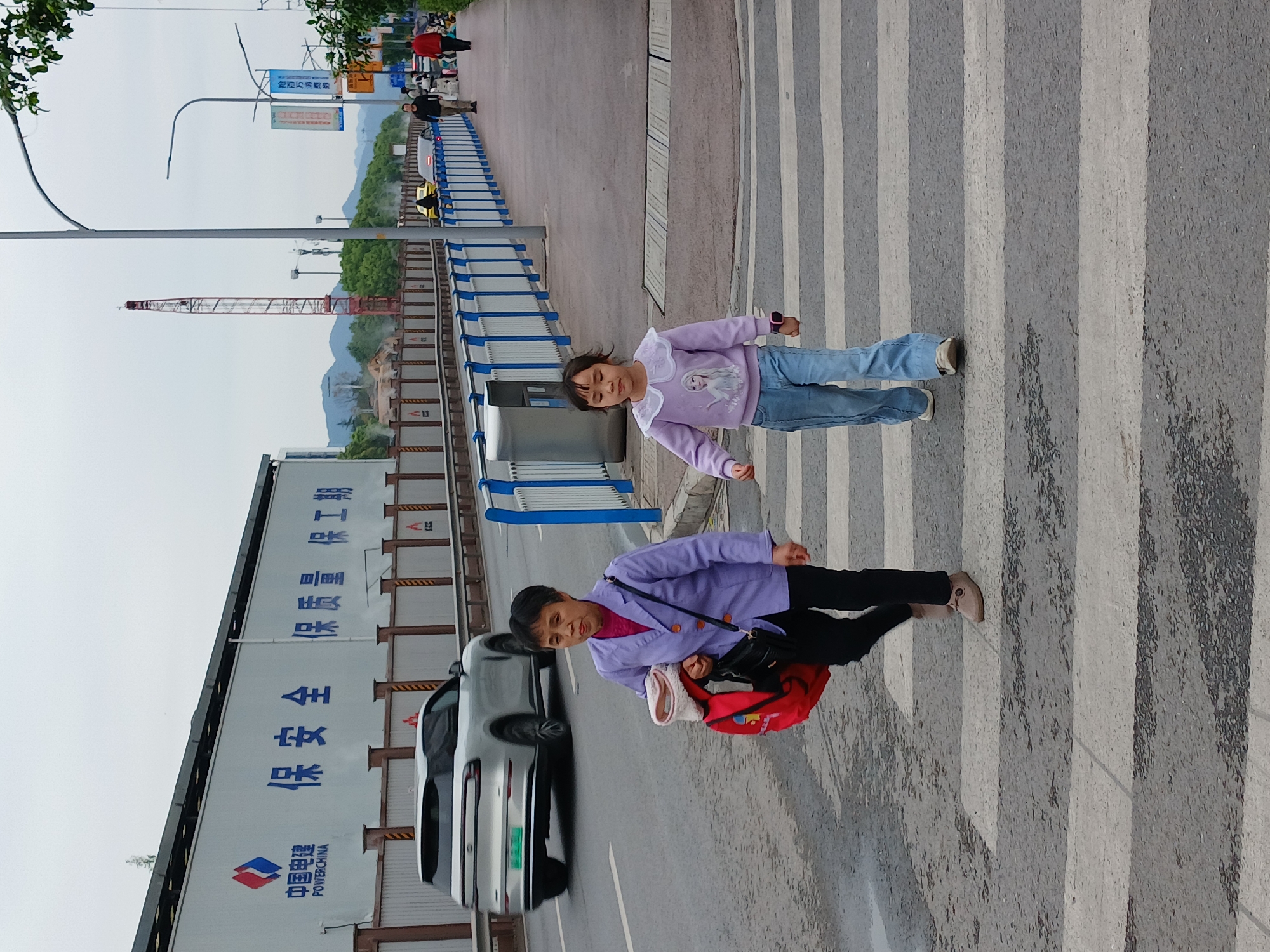 The image shows a woman and a young girl crossing a zebra crossing in what appears to be the Chongqing High-tech Zone in China. The woman is wearing a purple jacket and carrying a red bag, while the girl is wearing a light purple sweater and jeans. A white car is visible on the left side of the image. A construction site, which includes a billboard for POWERCHINA is also visible. The location is determined from the POWERCHINA sign on the billboard. A tall crane is visible on the background.