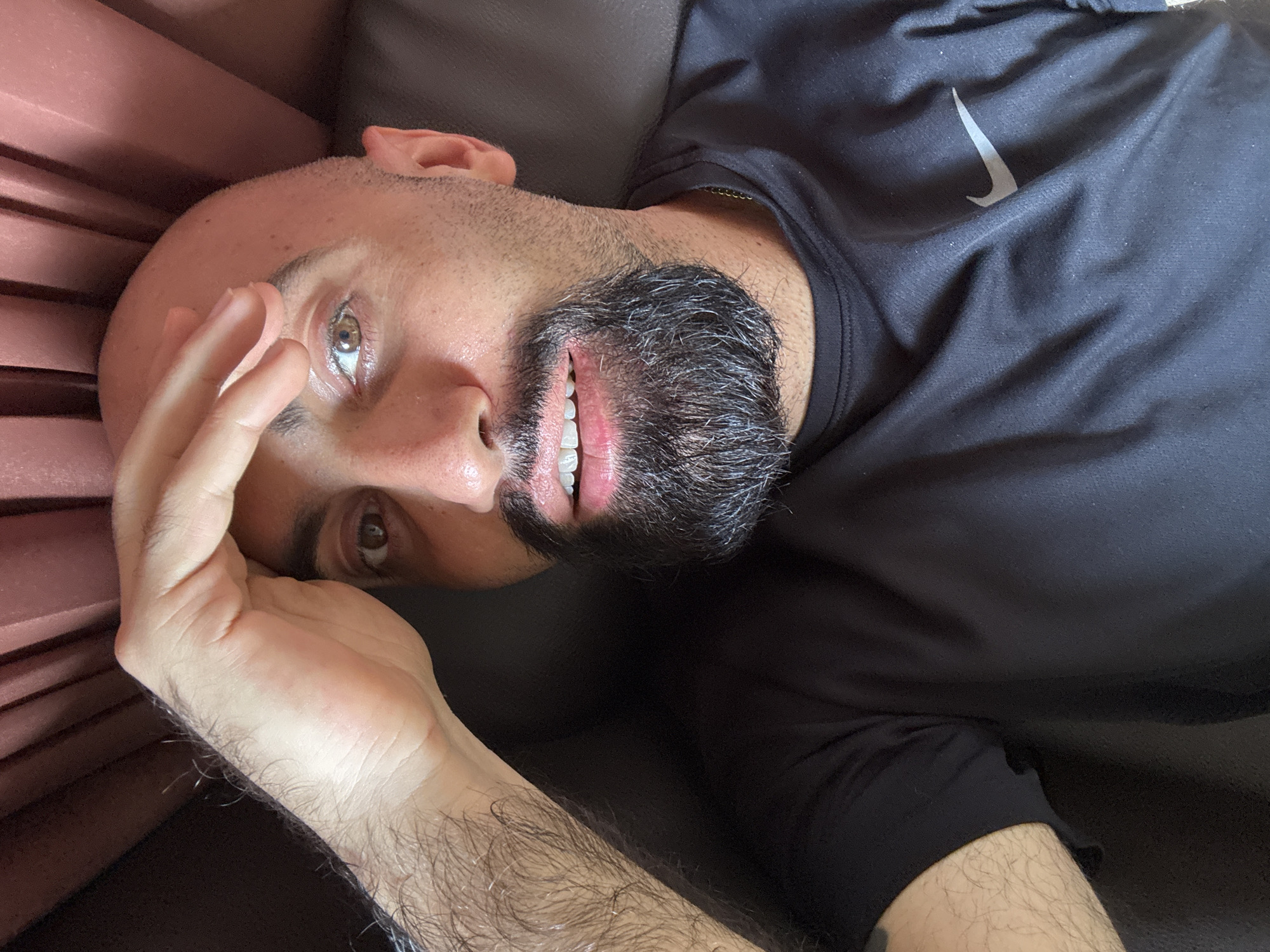 A bald man with a beard lies on a brown couch, propping himself up with his left hand on his forehead. He wears a black Nike t-shirt. The background is a blurry pink curtain and a brown couch. The man's left arm and chest are visible, as well as his right eye and mouth. The overall lighting is dim and natural, suggesting the photo was taken indoors during the day. There is no indication of the image being in Ko Samui District, Thailand.