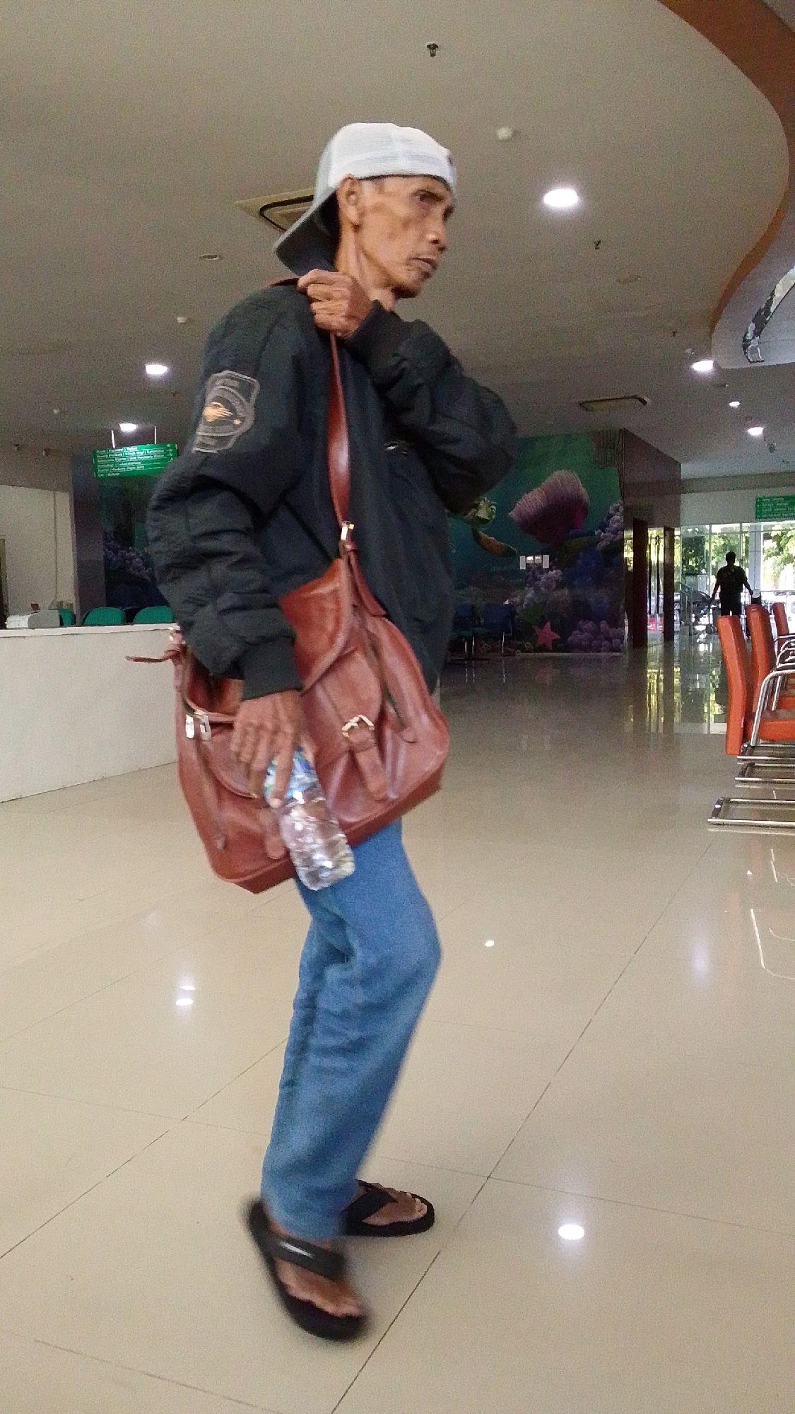 In a brightly lit indoor hallway with a polished tiled floor, an elderly Southeast Asian man walks from left to right. He is wearing a white baseball cap, a dark bomber jacket, blue jeans, and black flip-flops. A brown leather satchel is slung across his chest. He holds a clear plastic bottle of water in his right hand. In the background, there are rows of orange chairs, a reception desk, and a mural depicting an underwater scene. The hallway appears to be part of a public building, possibly a hospital or clinic, located in Pabelan, Indonesia.