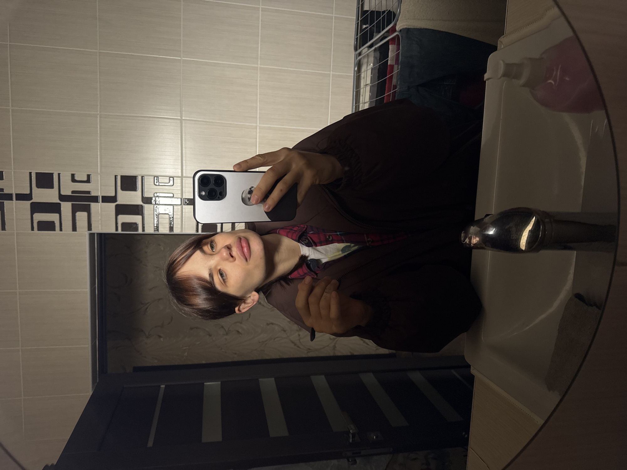 The image appears to be a selfie taken in a bathroom. A person is visible holding a smartphone to capture their reflection in a round mirror. The person has short, dark hair and is wearing a brown jacket over a red and white checkered shirt. The bathroom has beige and patterned tiles. A sink with a faucet and a soap dispenser are visible in the mirror's reflection. A partially visible metal rack with clothes on it can be seen in the background. There's no information in the image to verify the location in Rostov-on-Don, Russia.