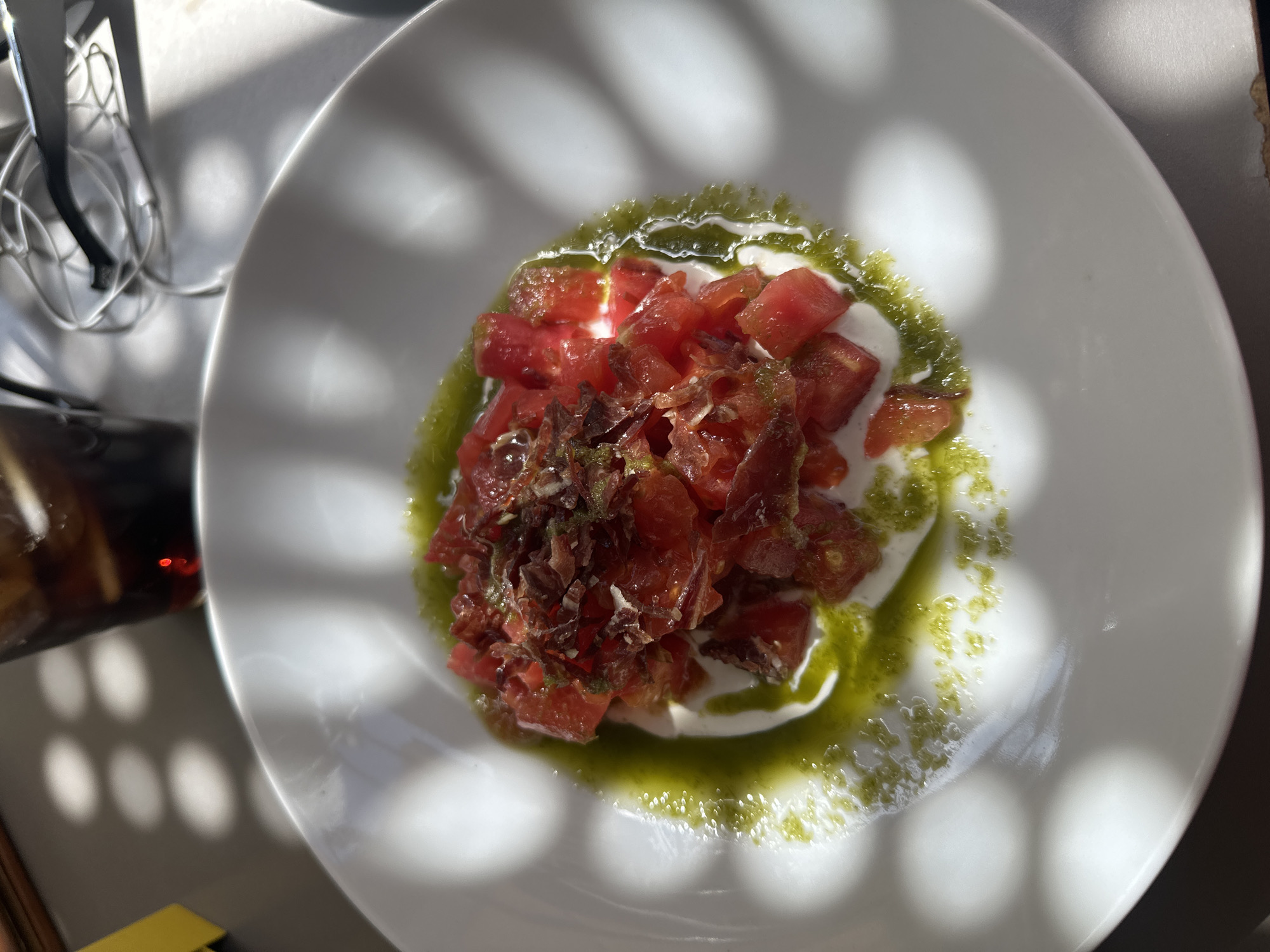 A bright, overhead shot captures a fresh dish served on a white, round plate, set on a light-colored table. The main subject is a culinary creation featuring diced red tomatoes atop a creamy white base, likely burrata or fresh mozzarella. A vibrant green oil or pesto is artistically drizzled around and over the ingredients. Scattered on top of the tomatoes are darker, shredded pieces, which appear to be cured ham, such as Jamón Serrano or Ibérico, a common delicacy in Madrid, Spain.
The scene is well-lit by strong, natural sunlight, casting distinct, circular and oval shadow patterns across the table and the plate. These shadows suggest a daytime setting, likely outdoors or by a window, with a patterned structure above. In the upper left corner, a metallic object, possibly part of a cutlery holder or a glass rack, is partially visible. The lower left of the frame shows a dark glass or bottle and a small yellow object, hinting at a broader table setting. The overall impression is one of a light, appetizing meal enjoyed in a sunny, pleasant environment. No people or visible text are present in the image.