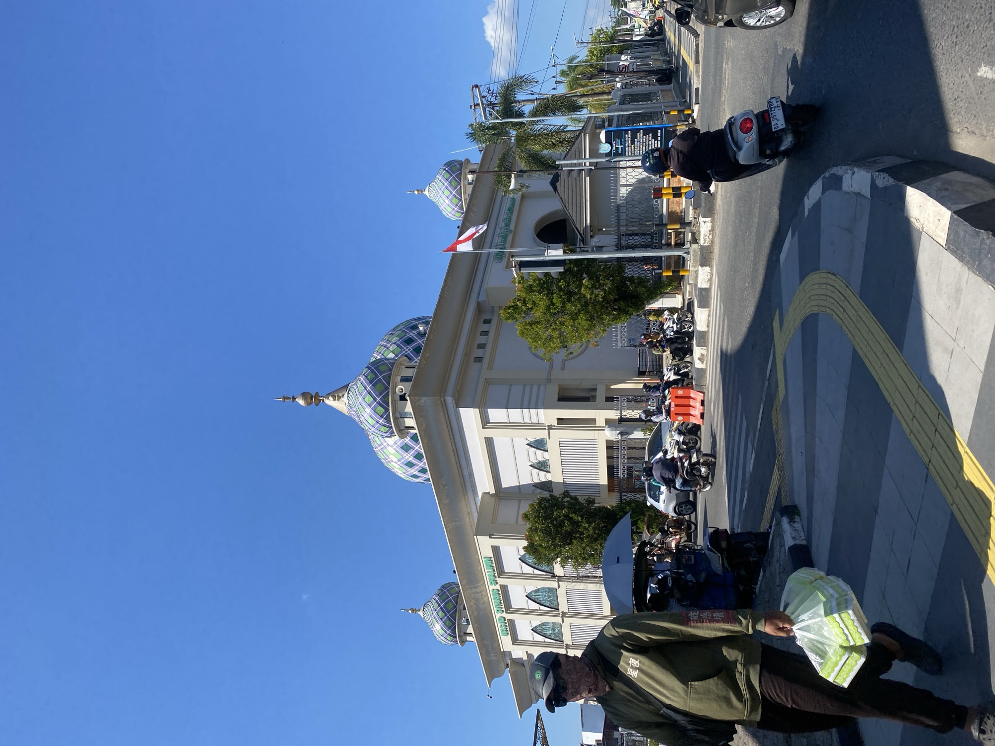 In the city of Jawa, Indonesia, under a clear blue sky, a mosque with intricate patterned domes stands prominently. The mosque, named "MASJID SYAHRIYAH SHALIM," features a white facade with decorative arches and windows. A red and white Indonesian flag flies from a flagpole outside the mosque. In the foreground, a person wearing a green hoodie, sunglasses, and a face covering walks with a plastic bag containing groceries. They are on a pedestrian crossing with yellow tactile paving. Several motorbikes and cars are present on the street, with some people riding motorbikes. Traffic cones and barriers are visible, suggesting ongoing construction or road work. Palm trees and other greenery surround the mosque, adding to the urban landscape. The scene depicts a typical day in an Indonesian city, with daily life and religious structures coexisting.