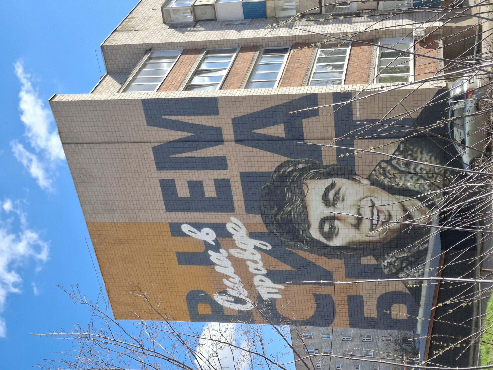 This image captures a large mural painted on the side of a multi-story building, set against a bright blue sky with sparse white clouds. The building features a facade of small, light-colored (beige/light brown) tiles and several rectangular windows, some with reddish-brown brick accents and external air conditioning units.
The mural depicts a prominent portrait of a smiling young man with dark, curly hair, identifiable as Danila Bagrov from the Russian film "Brother 2." The portrait is created with a mosaic-like effect using various shades of grey, black, and skin tones. Key text elements on the mural include the Russian phrase "СИЛА В ПРАВДЕ" (Strength in Truth) in stylized white script, and large dark blue/grey block letters forming "БРАТ 2" (Brother 2), set against a contrasting orange-yellow tiled background.
In the immediate foreground, numerous bare tree branches with nascent light green buds partially obscure the view, indicating springtime. The photograph is taken from a low angle, looking upwards, giving prominence to the sky and the upper sections of the building. The bright, even lighting suggests a sunny day, likely midday or afternoon. The scene portrays a piece of public art reflecting Russian culture within an urban or suburban environment in Sarapul, Russia.