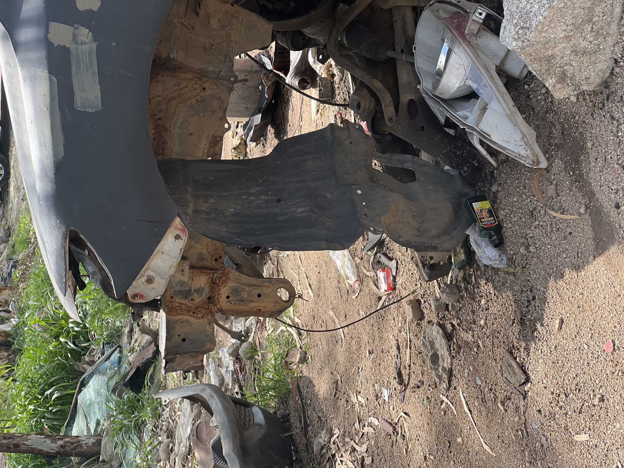 This image shows a close-up, angled view of damaged vehicle parts and debris scattered on the ground. The primary subject is the corroded and broken metal framework of what appears to be the front section of a car, including a fender, part of the chassis, and suspension components. A black plastic shield or undertray is also visible, as are pieces of a broken headlight assembly with a chrome reflector.
The scene is outdoors, with dry, sandy soil and small rocks covering the ground, interspersed with dry leaves and some sparse, dry vegetation. In the background, there is a hint of green grass and more debris, suggesting a junkyard or a disused area. The lighting indicates it is daytime, likely with bright sunlight casting shadows.
Among the debris, a dark green bottle with a label is visible, along with other plastic waste. The overall impression is one of disrepair and abandonment. There is no visible text that is clearly legible or significant to the scene. The image does not contain any people, and no specific event is taking place; it is a static depiction of damaged automotive components. The location cue provided is Howayi, Nigeria.