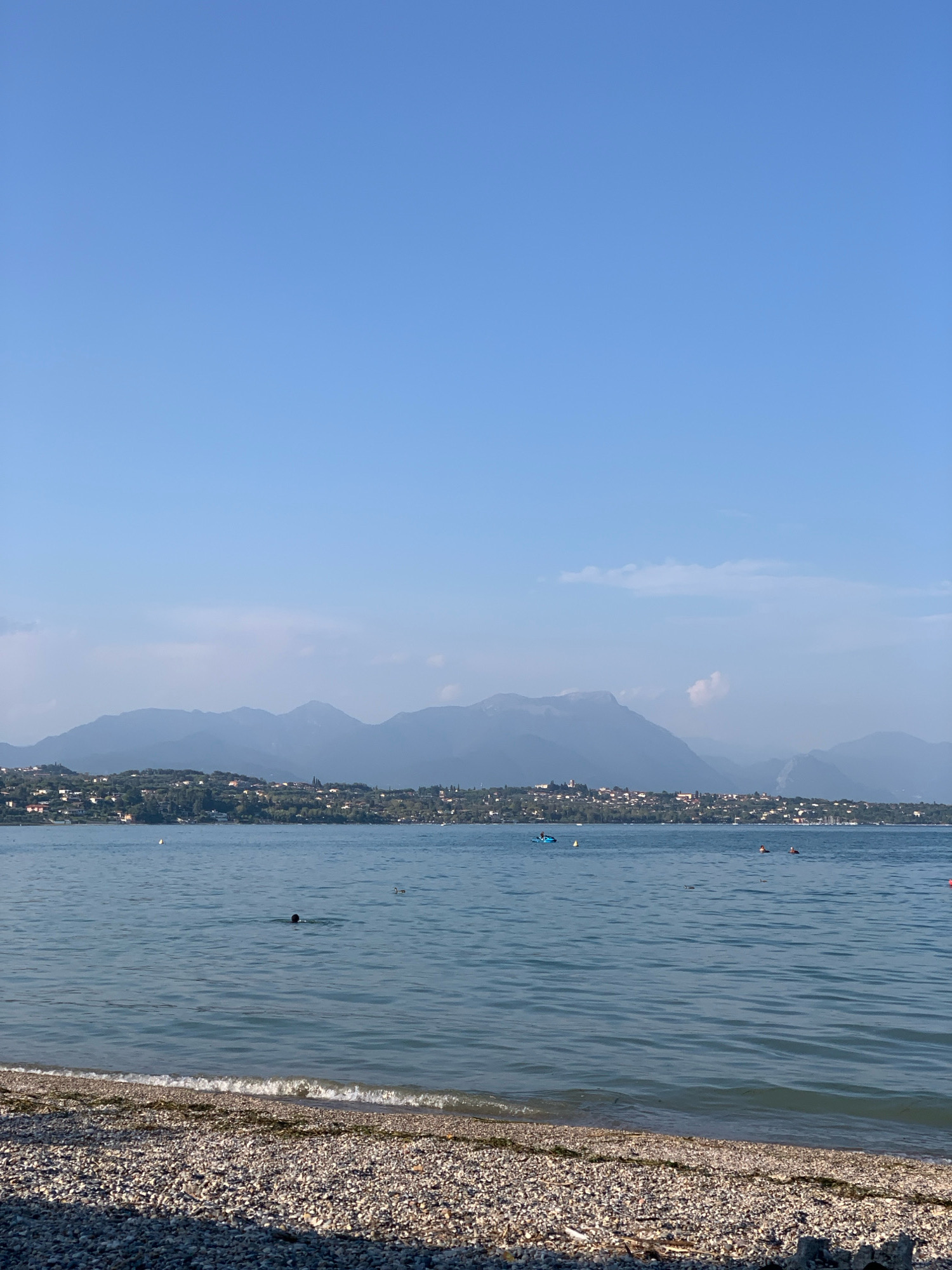The image shows a serene lake scene in Padenghe sul Garda, Italy. In the foreground, a pebble beach meets the calm, blue water of Lake Garda, with small waves gently lapping the shore. In the middle ground, several people are enjoying the water. One person is swimming with their head submerged, while others are visible as faint figures in the distance. A blue kayak or paddleboard is being used by someone further out on the lake. In the background, a picturesque town with red-tiled roofs is nestled along the shoreline, backed by rolling green hills and imposing, hazy mountains under a clear blue sky. The overall atmosphere is one of peaceful leisure on a sunny day.