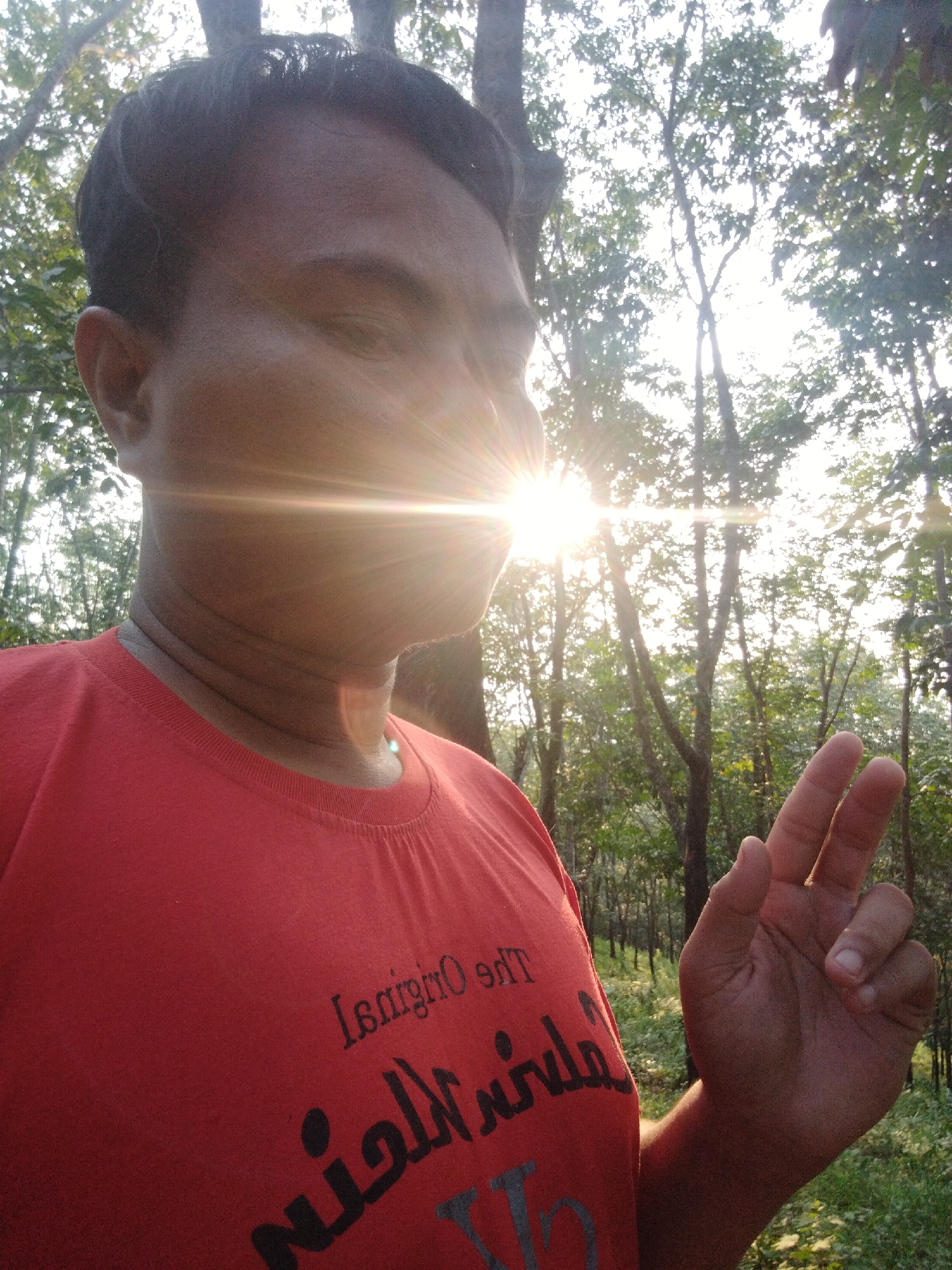 A man is standing outside in a forest area, holding up two fingers in a peace sign. He is wearing a red t-shirt with writing on it. The sun is behind his face, causing a glare and bright light rays. The location is Unknown, Unknown.