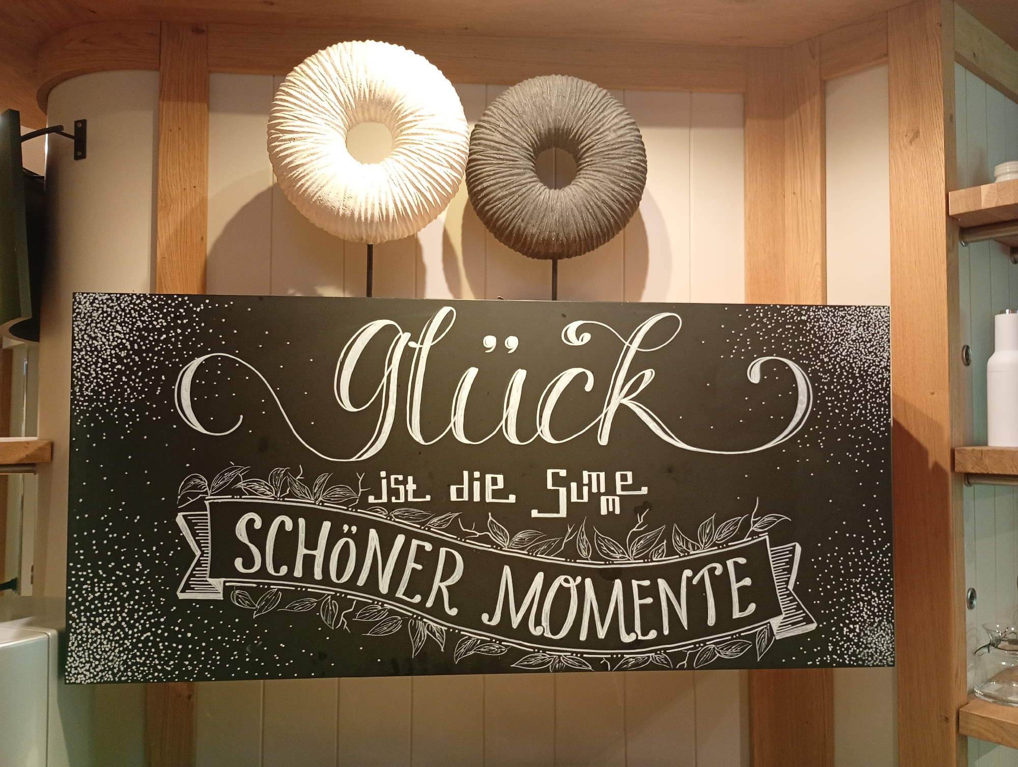 The image shows a sign with German text hanging on a black background. Above the sign are two round, ribbed decorative objects, one white and one gray, mounted on thin stands. The sign itself has a white, calligraphic inscription that reads "Glück ist die Summe Schöner Momente" which translates to "Happiness is the sum of beautiful moments." The text is embellished with leafy vines and a banner. Small white dots are scattered across the black background, resembling confetti or stars. The sign is displayed in a room with wooden beams and white paneled walls, suggesting a rustic or cozy interior. The background also includes glimpses of shelves with bottles and jars, hinting at a shop or cafe setting. Based on the provided context, this scene is located in Bad Sachsa, Germany.