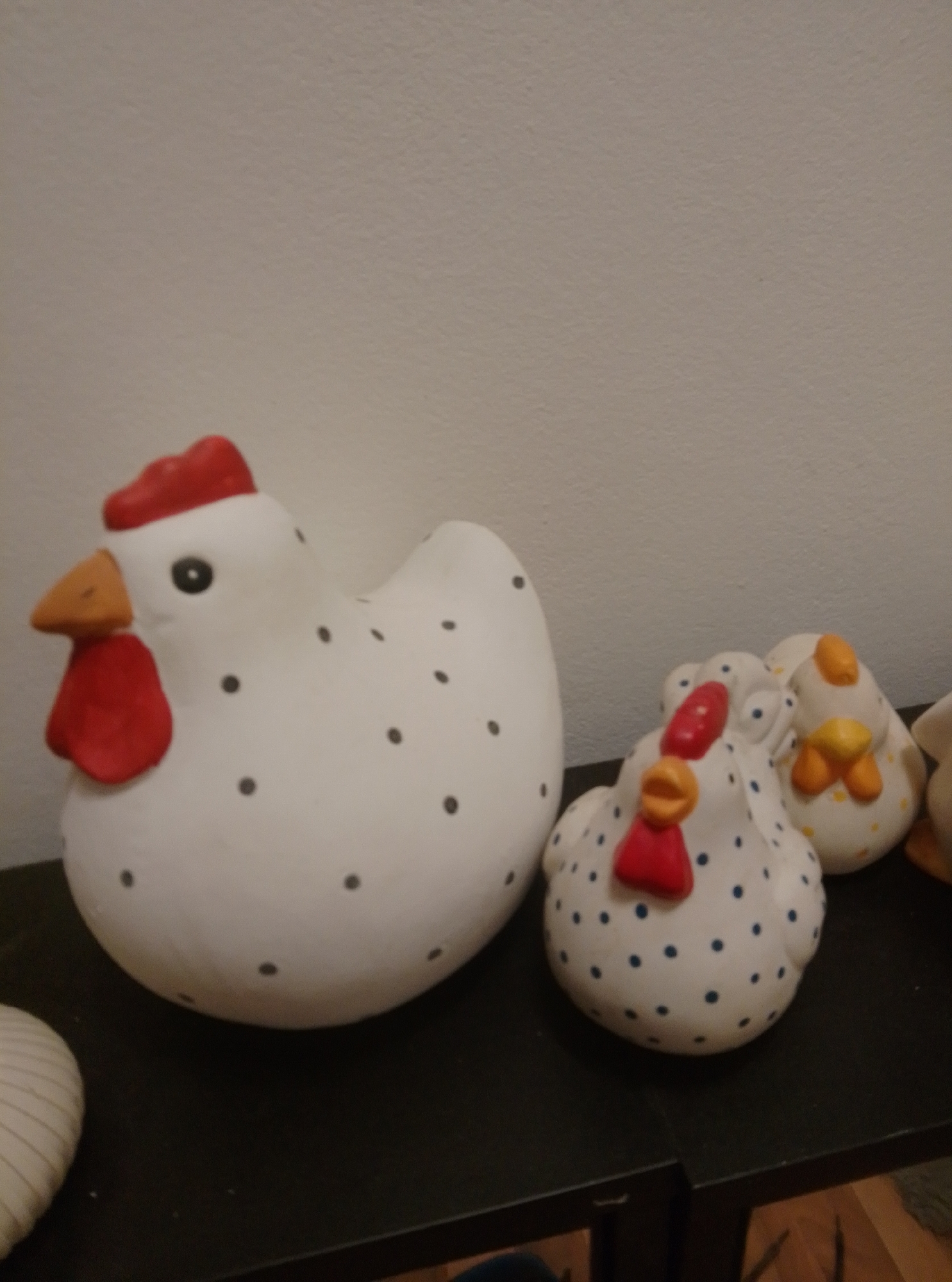 This image shows a collection of ceramic chicken figurines displayed on a dark surface, likely a shelf or tabletop, against a plain light-colored wall. The largest figurine, on the left, is white with black dots, a red comb, a yellow beak, and a red wattle. To its right are two smaller, similar figurines, also white with dots. The one in the middle has a red comb, yellow beak, and red wattle, while the one on the far right has a yellow comb, orange beak, and yellow wattles. Another white object with vertical stripes is partially visible on the far left. The overall lighting suggests an indoor setting. The image does not provide any specific information about the city of Wedel, Germany, or its context within the city.