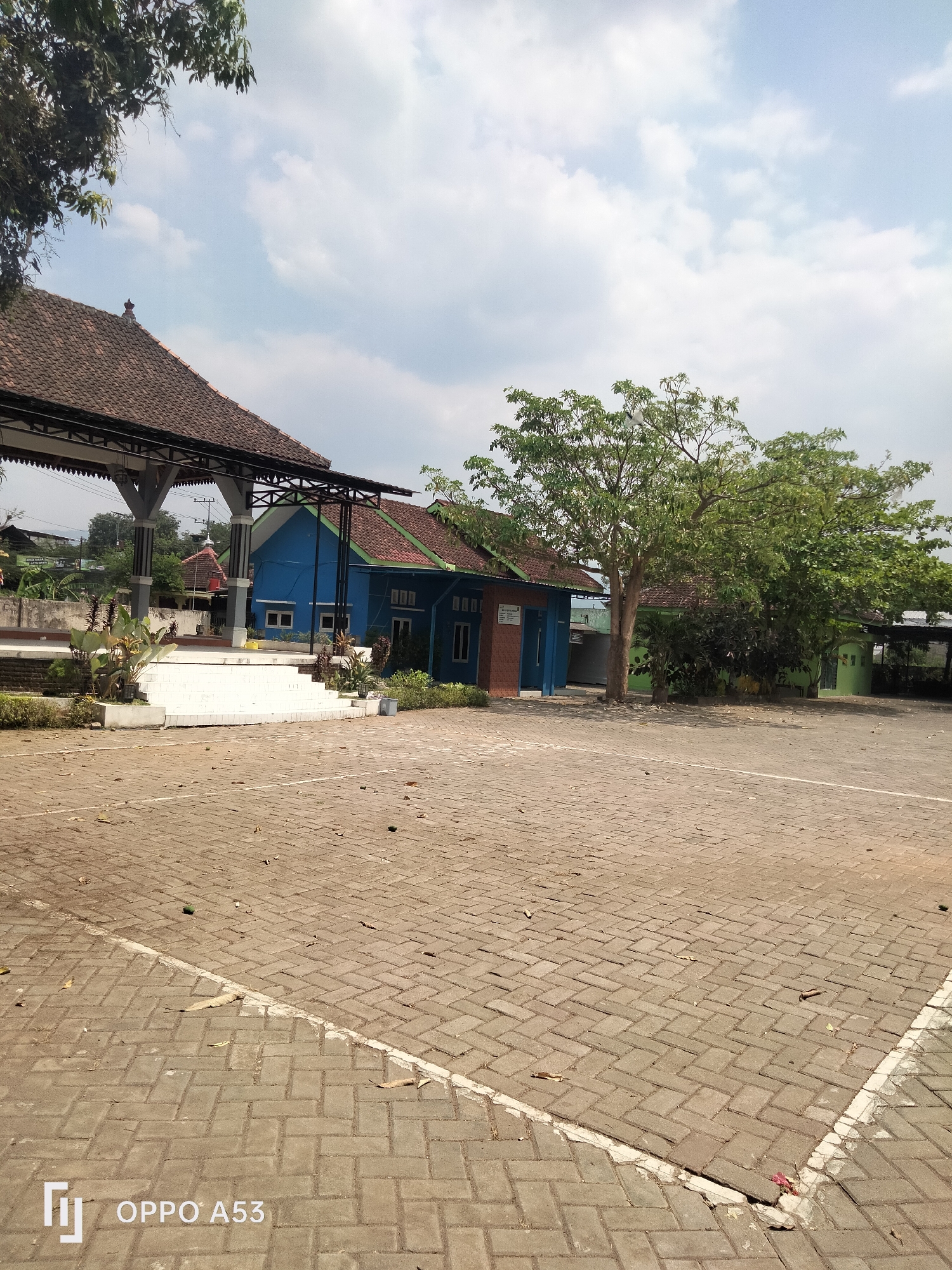 The image captures an outdoor scene with a paved courtyard in the foreground, composed of interlocking brick pavers. Scattered leaves and a few green objects, possibly fallen fruit, are visible on the pavement. A white line marks a boundary or parking space on the right side of the courtyard.
In the middle ground, a multi-tiered white stage or platform is present, bordered by planters with sparse vegetation. Behind the stage, a bright blue building with a red-tiled roof and visible windows stands prominently. To the right of the blue building, a smaller green building with a similar red-tiled roof is situated. Large trees with lush green foliage are scattered throughout the mid-ground and background, some casting shadows on the pavement. A dark-colored, open-sided structure with a tiled roof, likely a pavilion or shelter, is visible to the left, partially obscuring the view of the blue building. In the far background, other buildings and trees can be seen, suggesting a semi-urban or suburban setting. The sky above is partly cloudy with a light blue hue.
The watermark at the bottom left corner indicates the device used for the photograph: "LG OPPO A53". The overall scene appears to be a public space, possibly part of a school, community center, or religious complex in Karangan, Indonesia, given the context provided. No people are visible in the image.