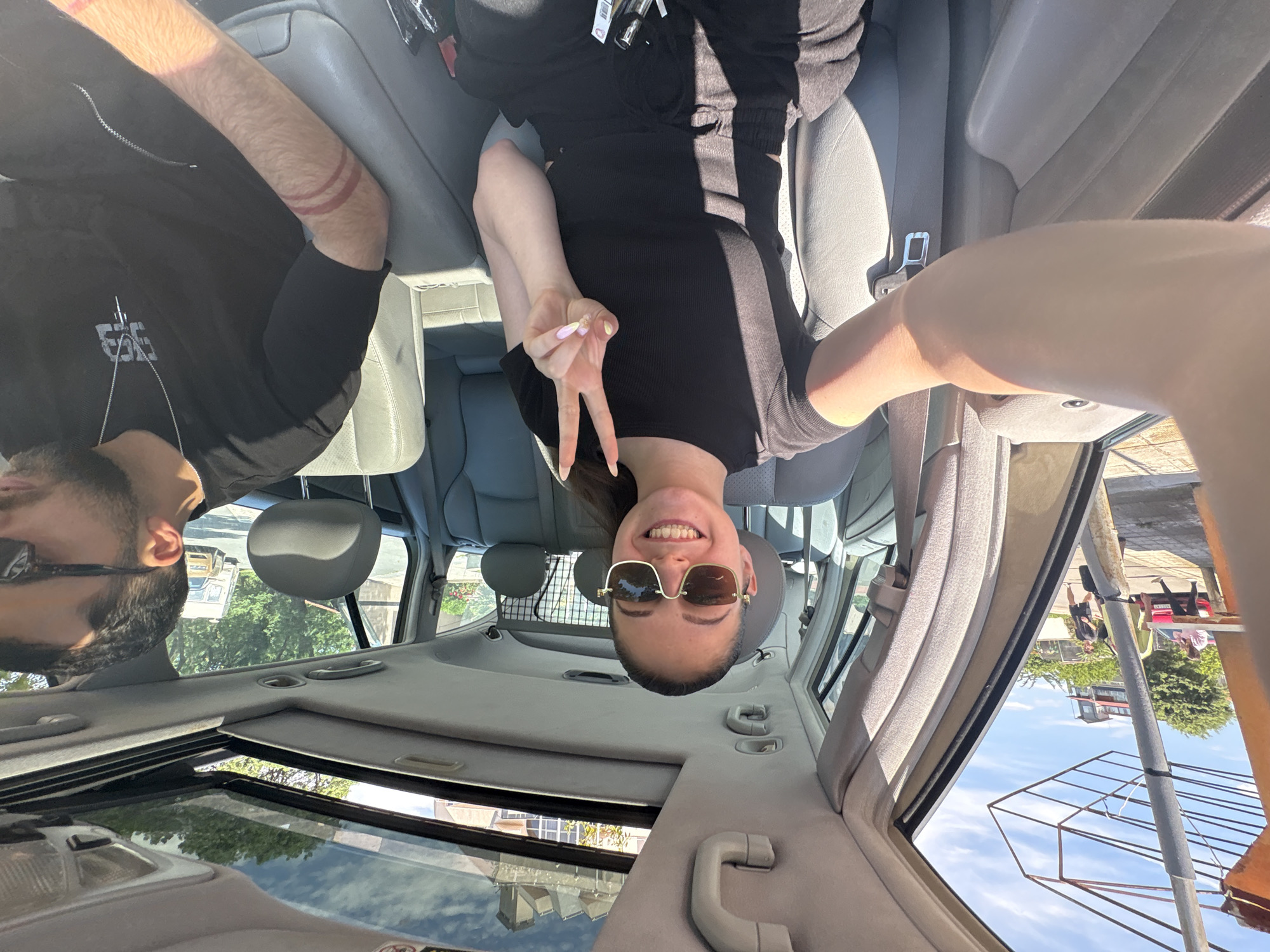 The interior of a car is visible, with a partially open sunroof. A woman wearing sunglasses smiles and makes a peace sign with her left hand. She is dressed in black. To her left, a man with a beard and sunglasses is seated. The car appears to be parked on a street in Tsarevo, Bulgaria, as suggested by a reflection in the sunroof which includes street objects such as buildings, vehicles, and street lights. The image is taken from an unusual angle, likely by someone in the car looking up toward the roof.