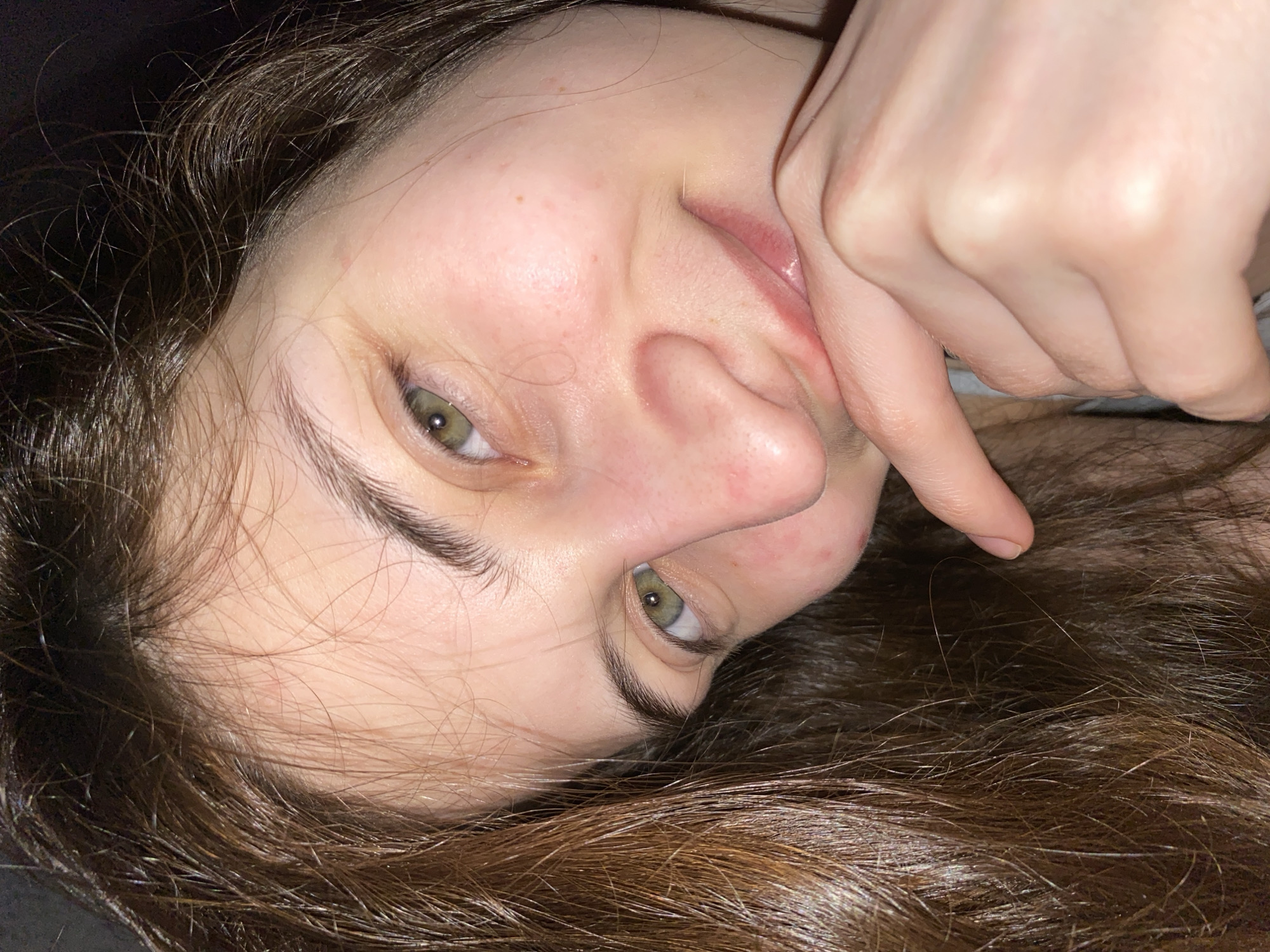 A close-up portrait features a young person, likely female, seen from an unusual, tilted perspective, possibly lying down. The subject has fair skin, prominent green or hazel eyes, and long, wavy dark brown hair that spreads around her head. Her face shows a natural appearance with some visible redness around the nose and cheeks. Her expression conveys a calm, perhaps playful, mood with a subtle smile or smirk on her lips. One hand, with fingers gently curled, is positioned near her mouth, with a finger lightly touching her lip or chin area. Her eyebrows are dark and well-defined. The background is uniformly dark and out of focus, suggesting an indoor setting, possibly at night or in a dimly lit environment, illuminated by artificial light. No text is visible in the image. The provided location context is Votkinsk, Russia.