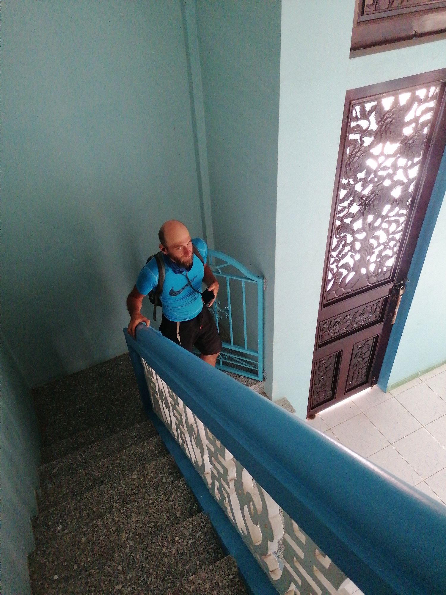 A bald, bearded man wearing a blue t-shirt, black shorts, and a black backpack walks up a granite staircase. He is holding a black smartphone in his right hand and has earbuds in his ears. The staircase has a blue railing with a decorative white insert. The walls are painted a light blue. To the right of the staircase, there is a brown wooden door with intricate carvings and a light blue metal gate. The scene appears to be an interior staircase in Lộc Quý, Vietnam.