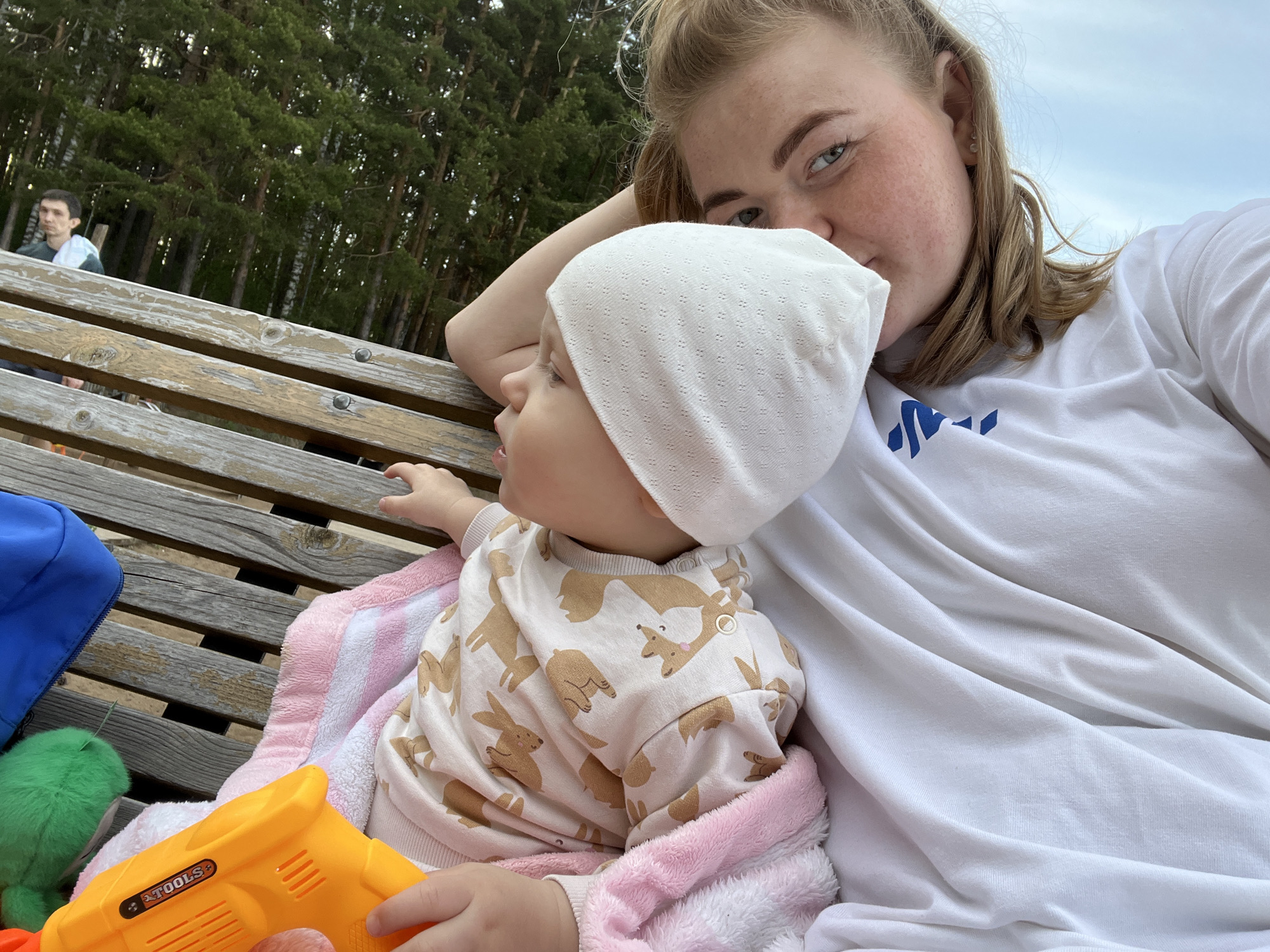The image shows a fair-skinned woman with reddish-blonde hair and freckles posing for a selfie with a baby, presumably in Chaikovsky, Russia. The woman is wearing a white t-shirt with blue markings on the chest. The baby is fair-skinned and wearing a white hat and a white and tan patterned top with images of animals. They are sitting on a wooden bench. A pink and white blanket is beneath the baby. A blue backpack, a green stuffed animal, and a bright orange plastic toy are next to them on the bench. In the background, out of focus, are trees and a partially visible person wearing a white towel draped over their shoulder. The sky is visible in the upper right corner.