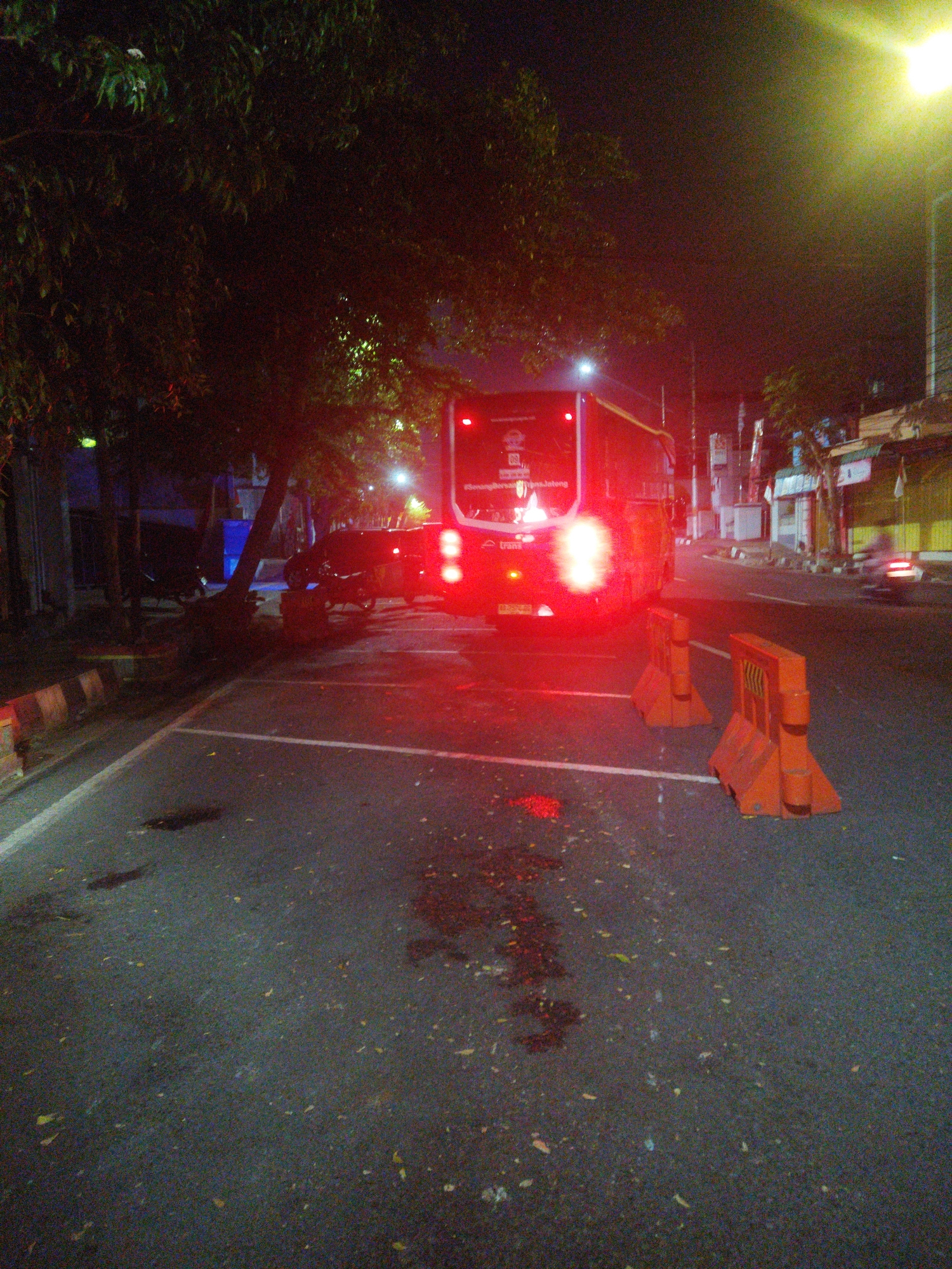 The image captures a nighttime scene on a street in Wonogiri, Indonesia. A large red bus is parked on the right side of the road, with its brake lights illuminated. Two orange traffic barriers are positioned in front of the bus, one slightly closer to the viewer than the other. The street is paved and appears wet in places, with some scattered leaves on the asphalt. Trees with dense foliage are visible on the left side of the image, casting shadows and partially obscuring the view of buildings and other parked vehicles behind them. A street light with a bright yellow glow is visible in the upper right corner, illuminating the surrounding darkness. The overall atmosphere is dim, with the red light from the bus's taillights being the most prominent source of illumination.