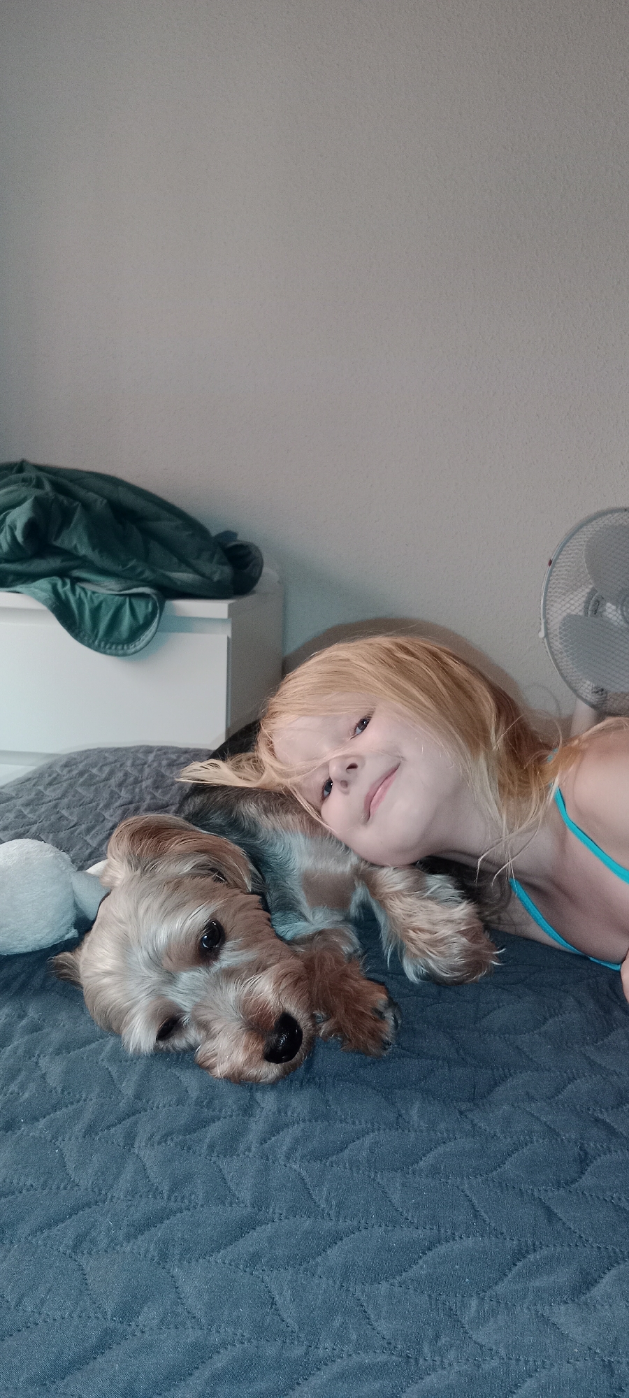 A young girl with blonde hair and a turquoise top lies on a bed next to a small, light brown dog. The girl is resting her head near the dog, and both are looking towards the camera with relaxed expressions. The dog's head is resting on the girl's arm. They are on a dark grey quilted bedspread. In the background, to the left, is a white chest of drawers with a folded green blanket on top. To the right, a white oscillating fan is visible against a pale grey wall. The lighting suggests it is daytime. The overall mood is peaceful and affectionate. There is no visible text.