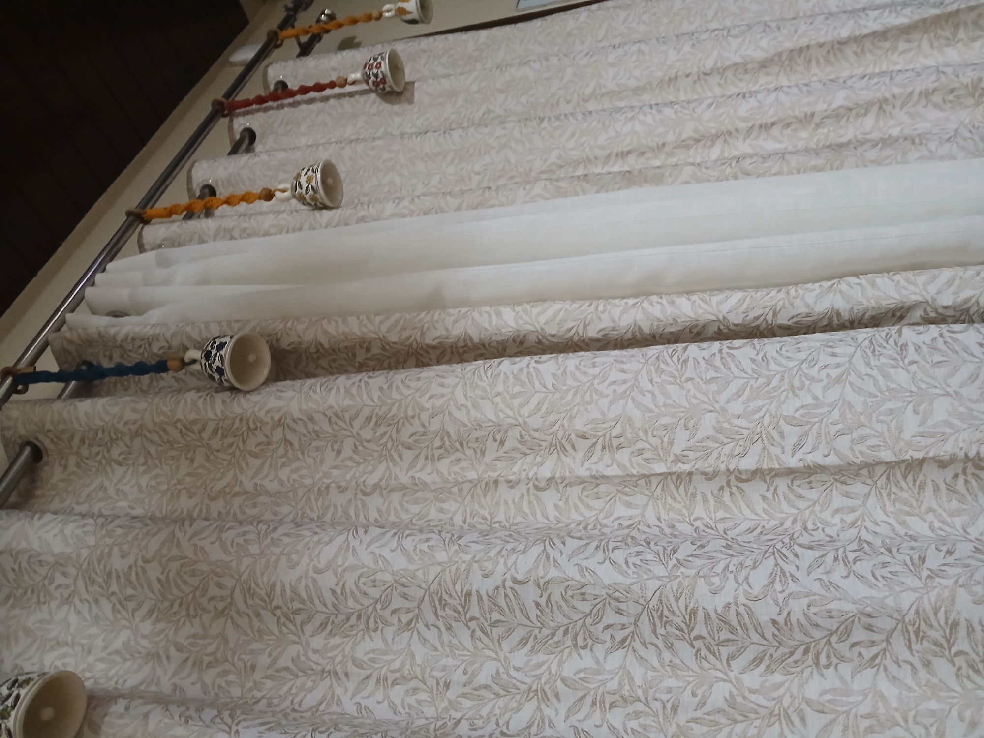 The image depicts a curtain rod holding up a multi-layered curtain. The curtain consists of a patterned fabric, likely a floral or leaf design in neutral tones, and a white, sheer fabric layer. The curtain rod is a metal pole affixed to the wall with brackets. Decorative ceramic or porcelain bells are suspended from the rod by cords or strings between the brackets and the curtain. The setting appears to be indoors, possibly a residential space. There is no specific information to determine if it is Bengaluru, India.