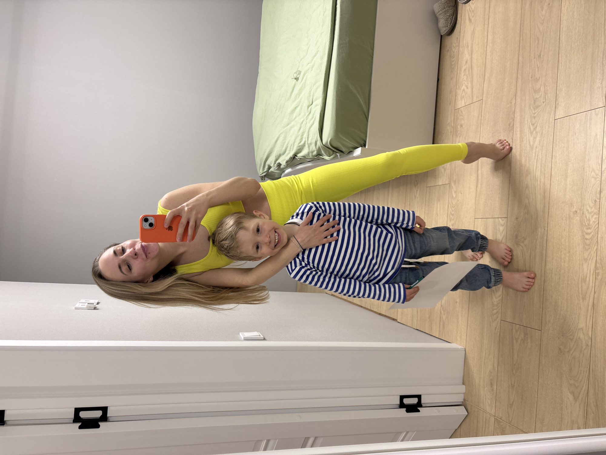 An indoor selfie captures a smiling woman and a young boy, appearing to be mother and son, in a home setting in Chaikovsky, Russia. The woman, with long blonde hair, is wearing a bright neon yellow ribbed athletic top and matching leggings. She holds an orange smartphone, identifiable by an Apple logo, to take the picture, extending her arm around the boy. The boy, also with blonde hair, is dressed in a blue and white striped long-sleeve shirt and blue jeans. He is barefoot and holds a white piece of paper in his left hand. Both subjects are smiling broadly at the camera, conveying happiness and affection. The background features a light gray wall, a light wooden floor, and a light green bed with a wrinkled duvet. A white door frame and a light switch are visible on the left side of the frame. A pair of grey slippers are also visible on the floor near the bed. The scene suggests a cheerful, intimate moment captured during the daytime.