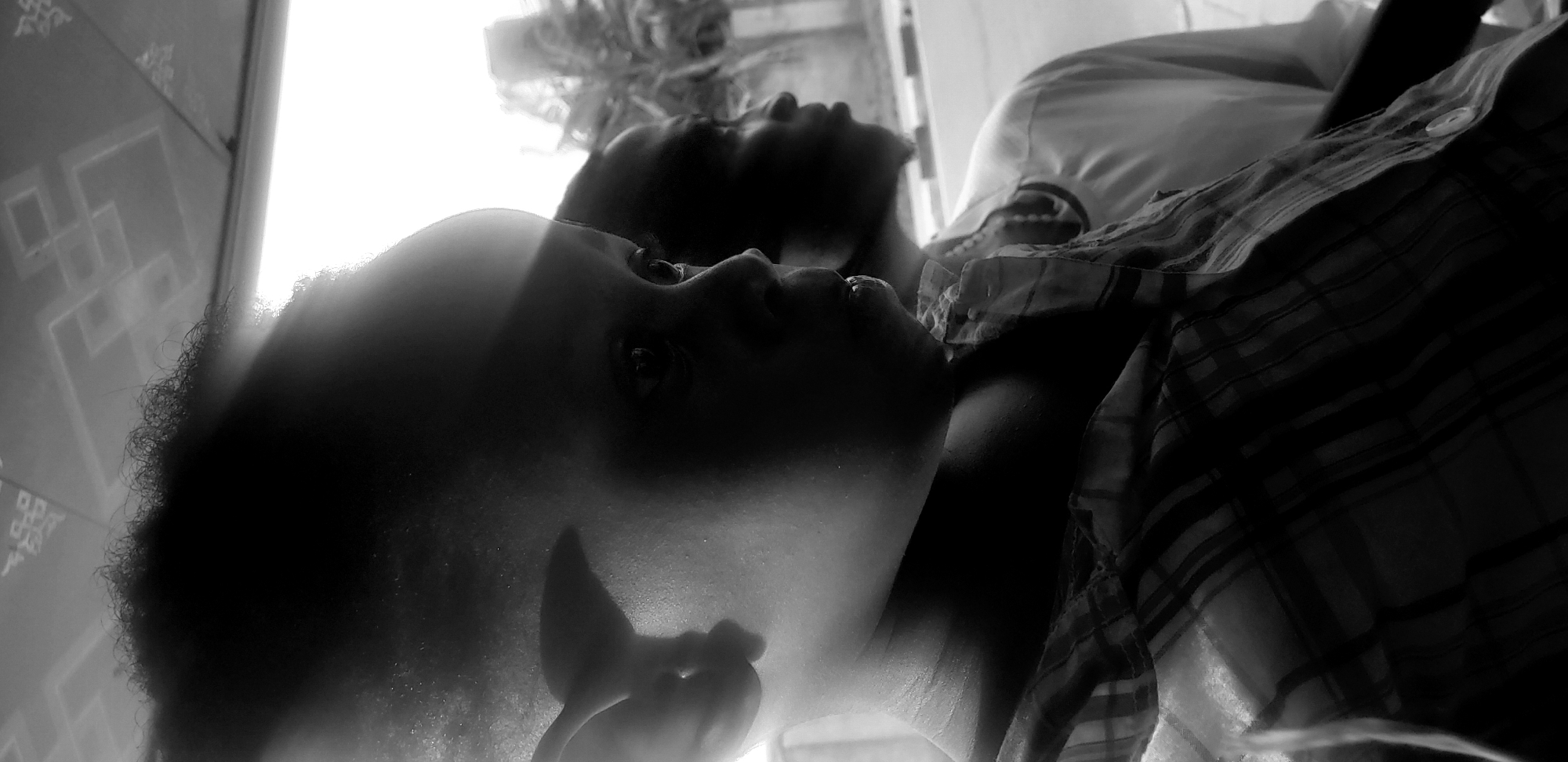 This black and white media file captures a close-up, angled shot of two individuals lying down, their faces tilted upwards towards a bright light source. The subjects appear to be young. The person in the foreground is closer to the camera, with their face partially obscured by shadow. Their eyes are open and looking upwards, and their curly hair is visible. The second person is positioned behind and slightly to the side of the first, their face also directed upwards. They are wearing a collared, plaid shirt, a button of which is visible. The background consists of a textured surface, possibly a wall or patterned fabric, with geometric shapes. The lighting is strong and backlit, creating high contrast and obscuring some details. The scene suggests a casual, indoor setting, possibly a bedroom or living area, in Narayi, Nigeria. The overall mood is introspective or contemplative due to the upward gaze of the subjects. There is no visible text in the image.